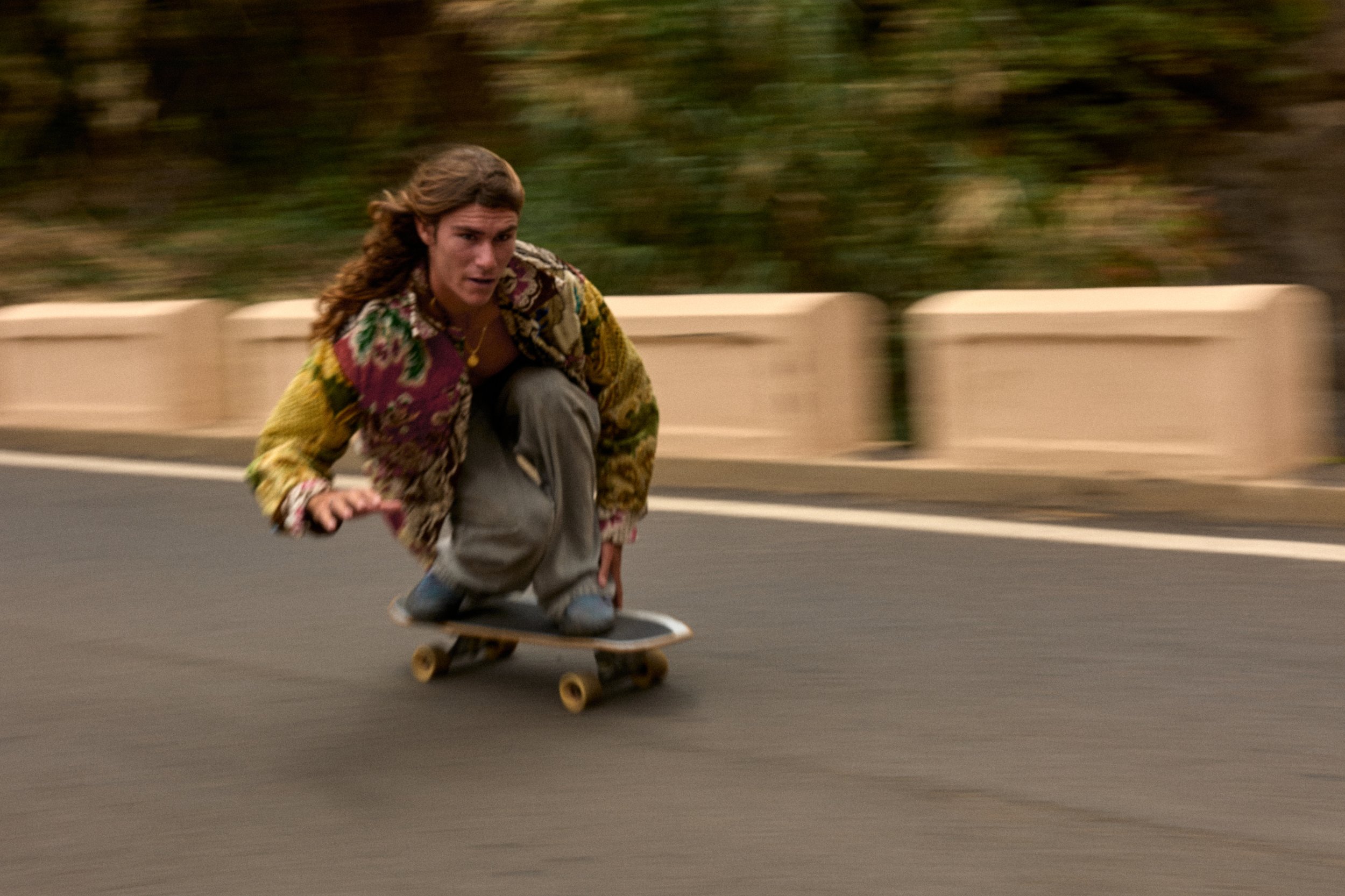 A person with long hair wearing a multicolored jacket and gray pants skateboarding down a road.