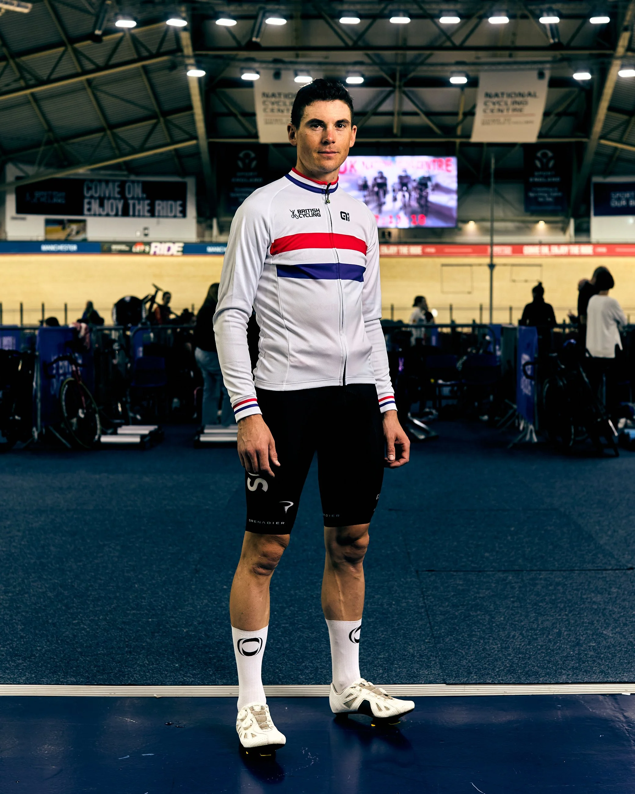 A male cyclist stands in an indoor velodrome, wearing a white cycling jersey with red, white, and blue stripes, black cycling shorts, white compression socks, and cycling shoes. The velodrome has a high arched ceiling, with bicycles and people in the