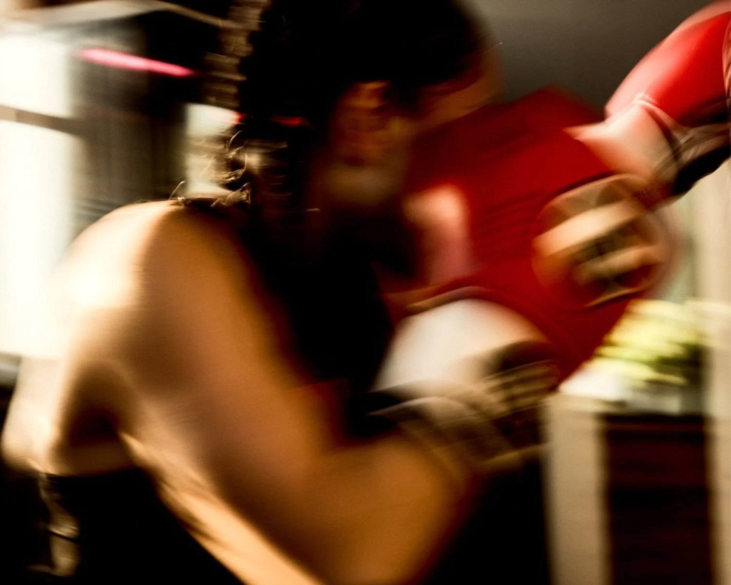 A blurred person with dark hair playing an acoustic guitar, wearing a red shirt, in a dimly lit room.