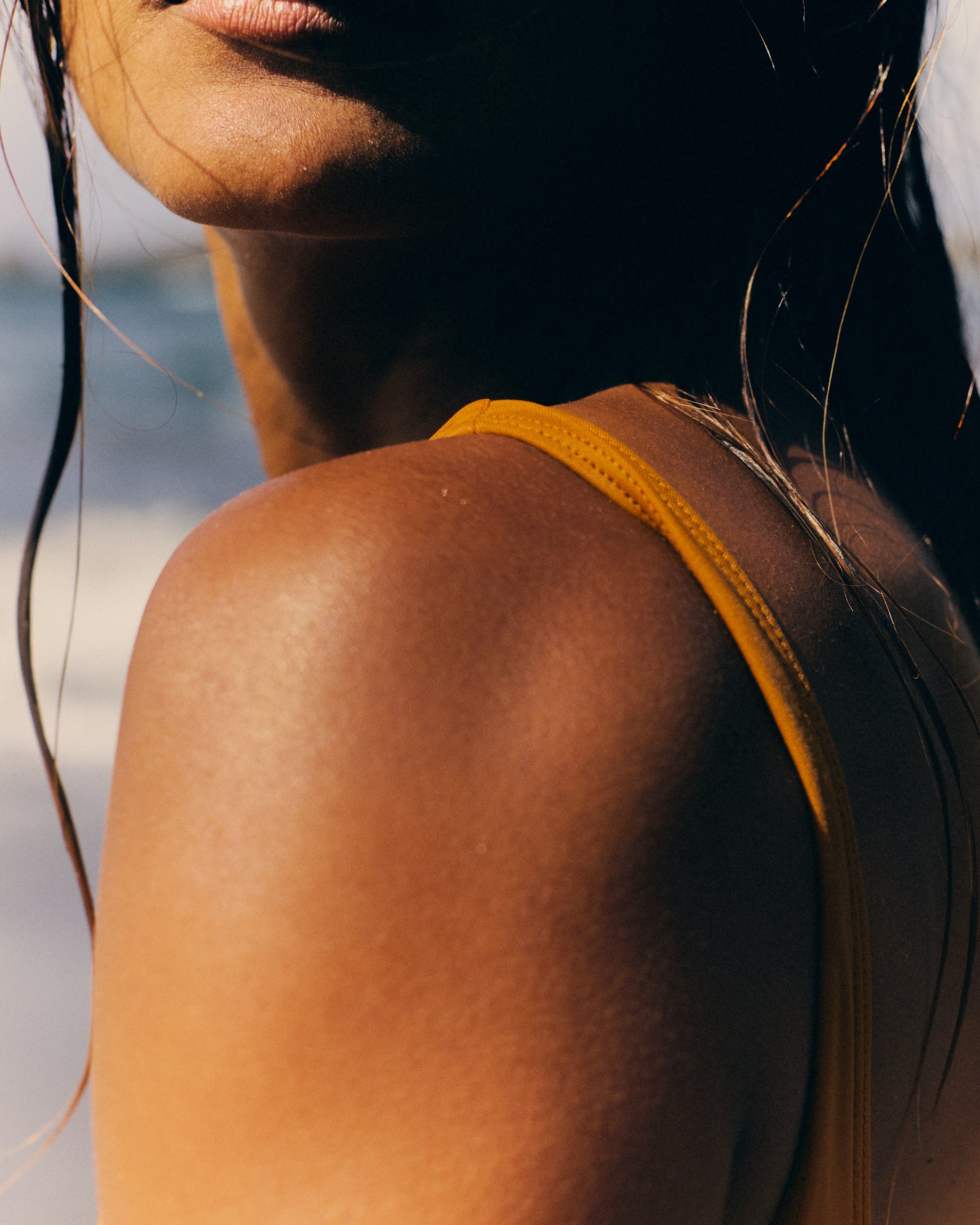 Close-up of a woman's shoulder and part of her face, wearing a yellow swimsuit, with sunlight highlighting her skin.