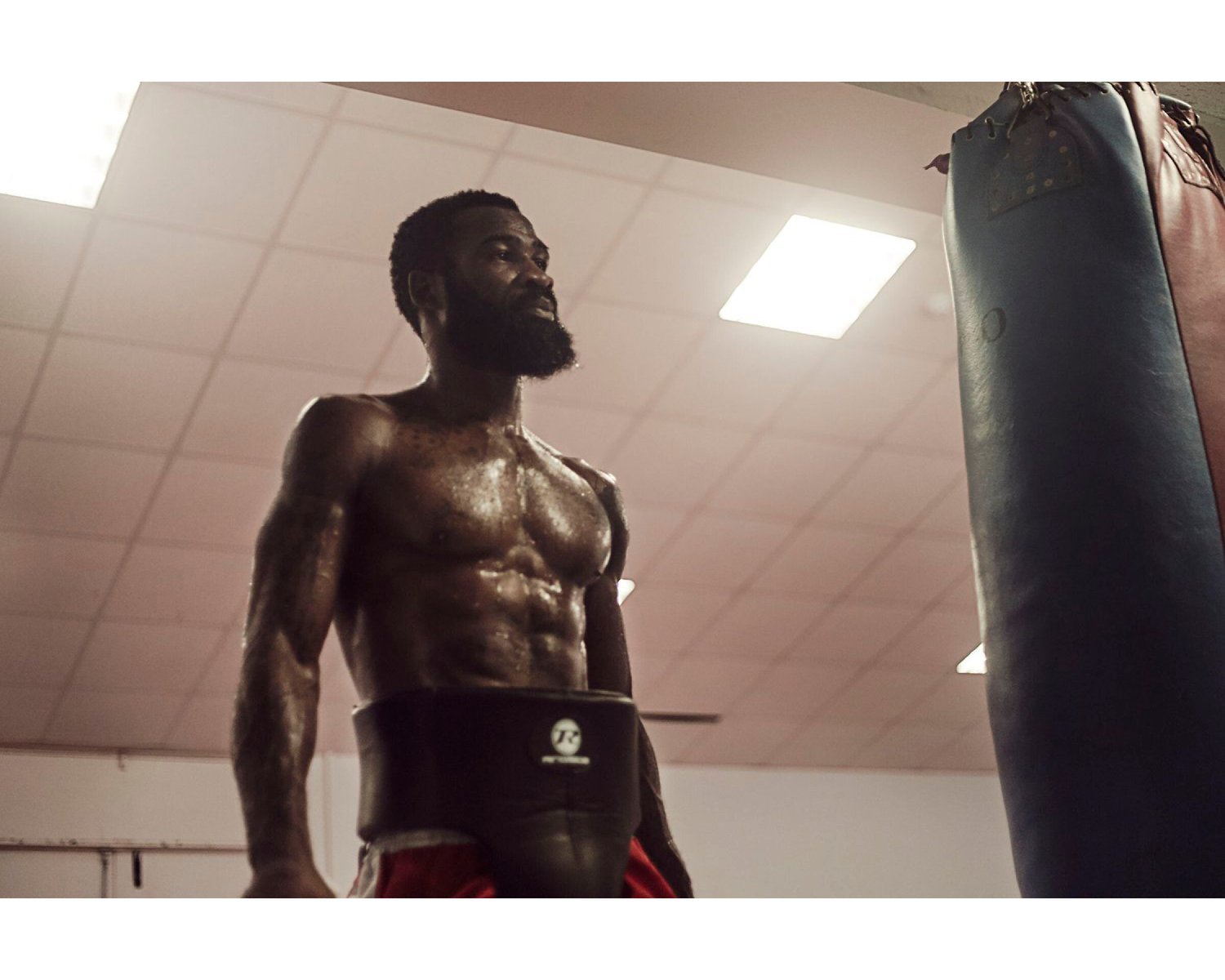 A shirtless man with a beard and tattoos on his arms standing near a punching bag in a gym.