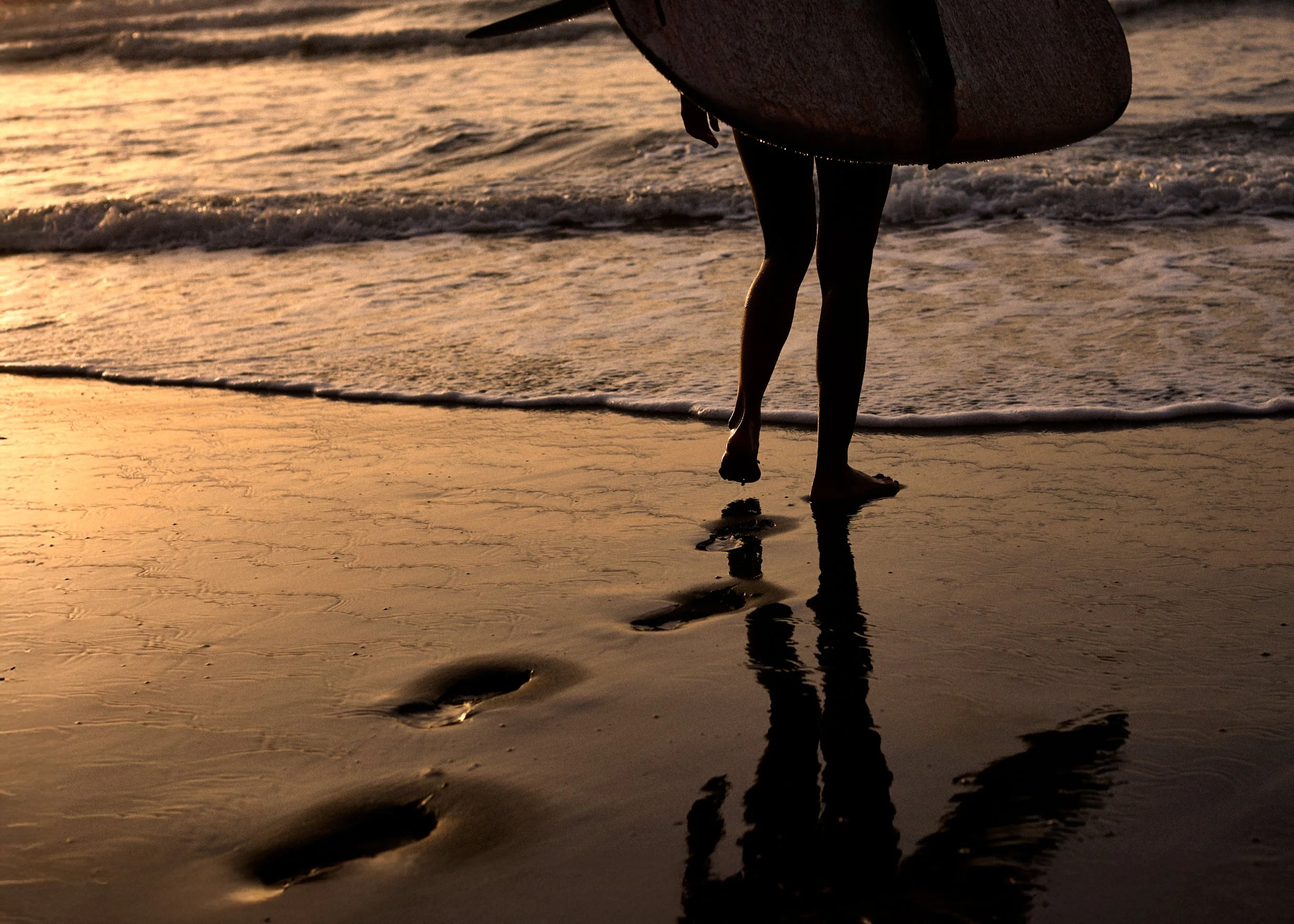 A person walking along the beach at sunset, carrying a surfboard under their arm, with footprints in the wet sand reflected in the water.
