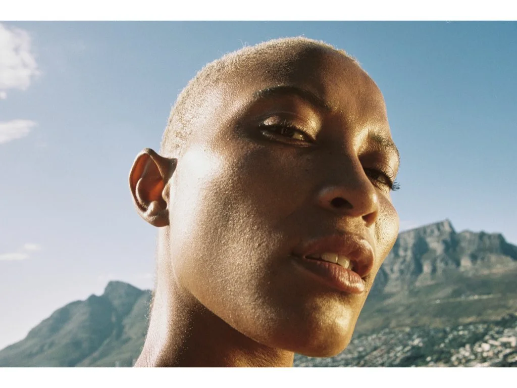 Close-up of a person's face with short blond hair, gold eyeshadow, and natural makeup outdoors with a mountain in the background during daylight.