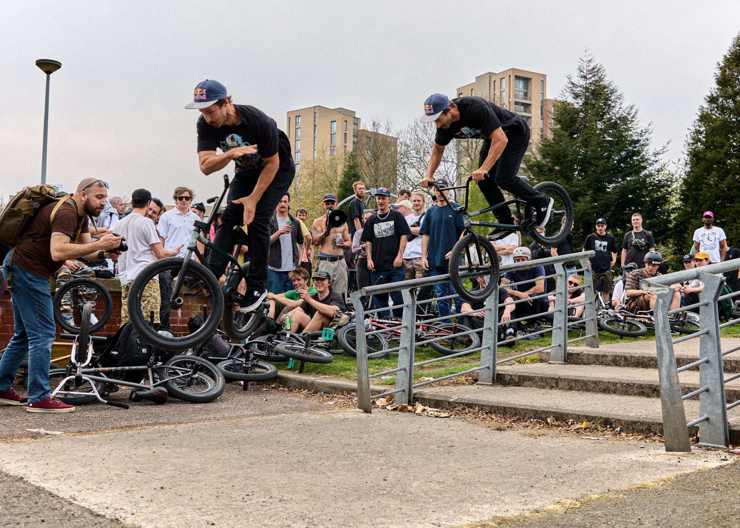 BMX bikers jump over a rail during a competition while a crowd of spectators and photographers watch nearby.