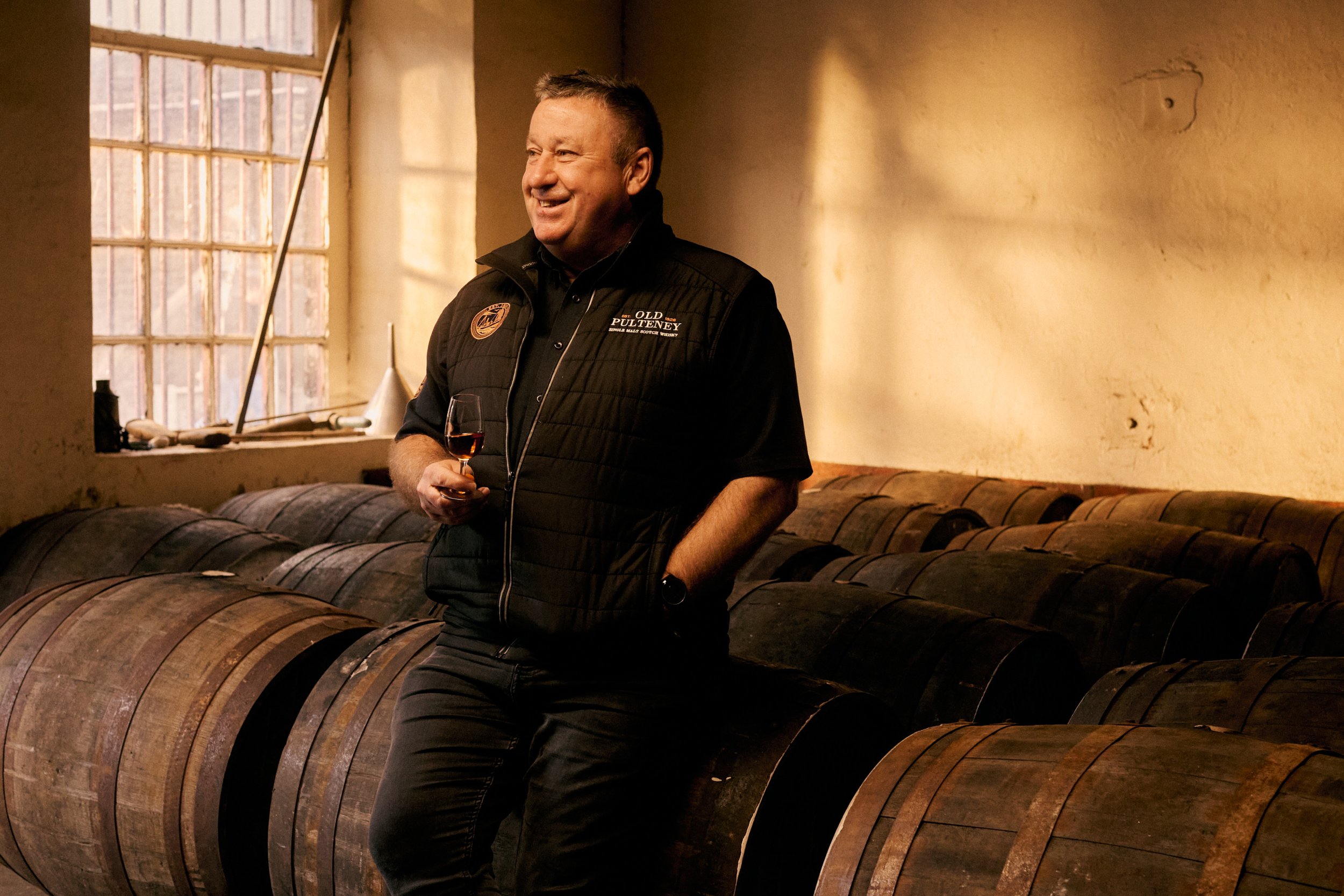 A man in a black vest and black shirt holding a glass of red wine, standing in a room with a rustic, cozy atmosphere, surrounded by wooden barrels and a large window letting in natural light.
