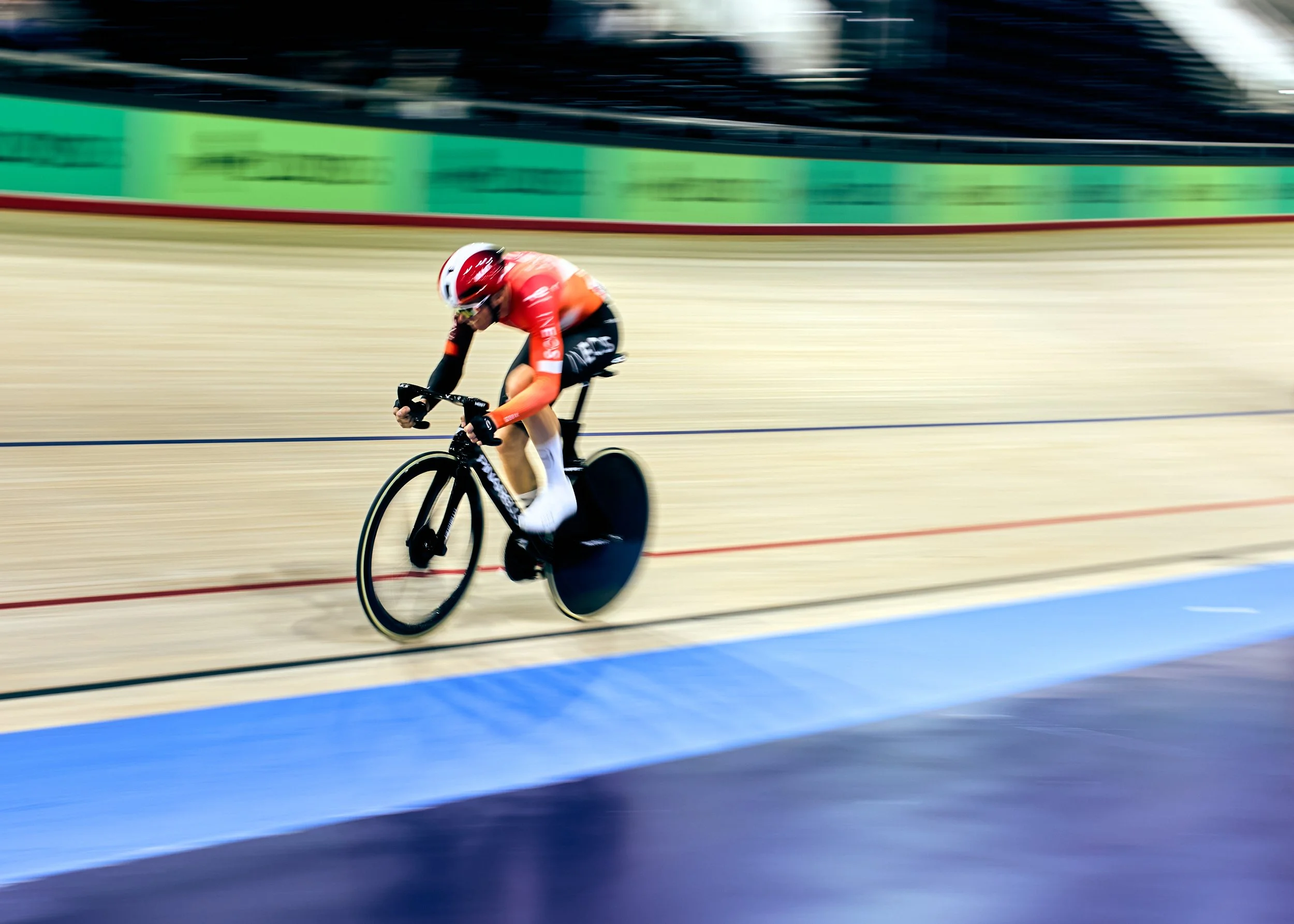 A cyclist wearing a red helmet and orange jersey riding on an indoor velodrome track with blurred background indicating high speed.