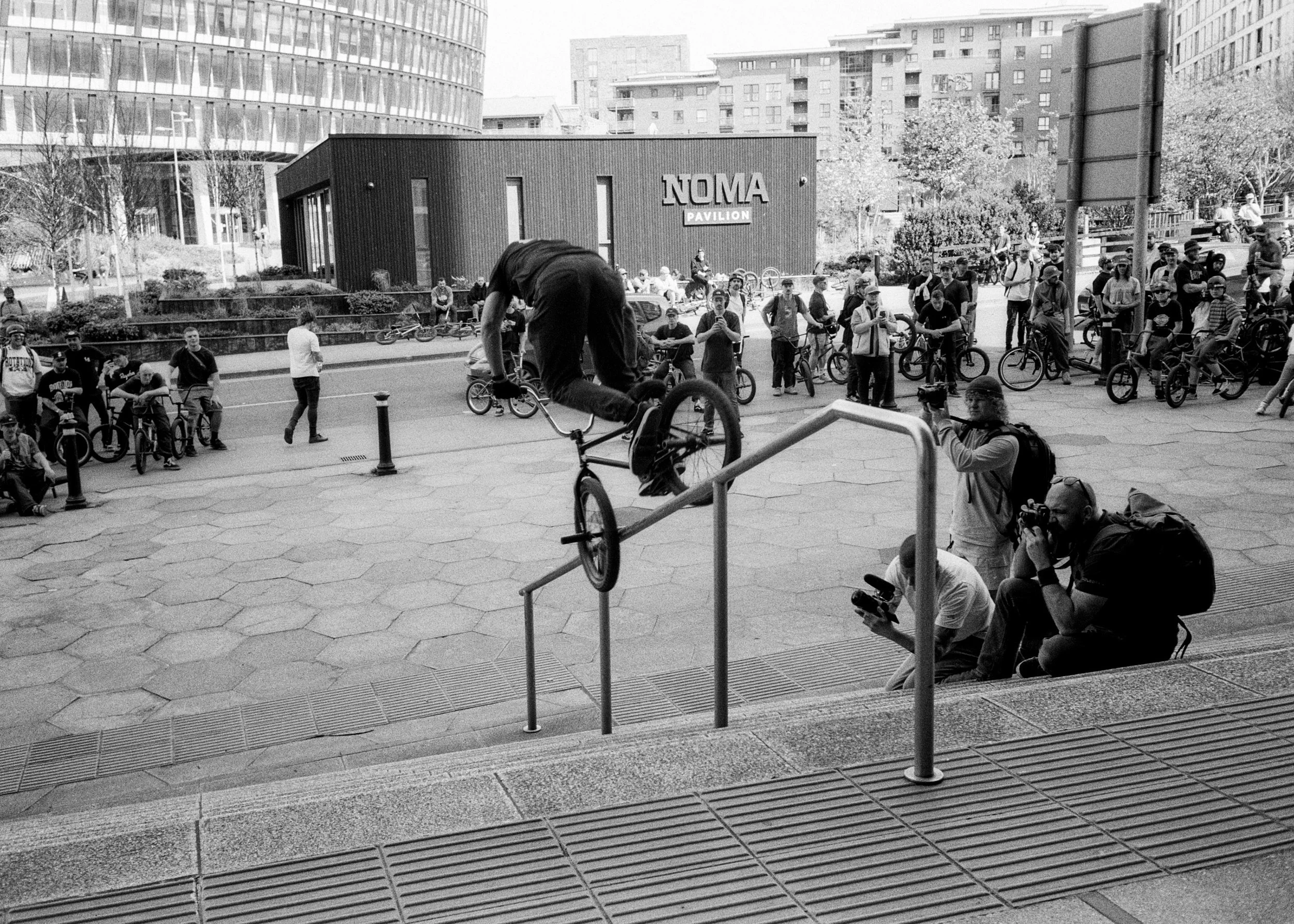 A person performing a BMX stunt on a handrail while a crowd of onlookers and photographers watch in an urban plaza with modern buildings in the background.