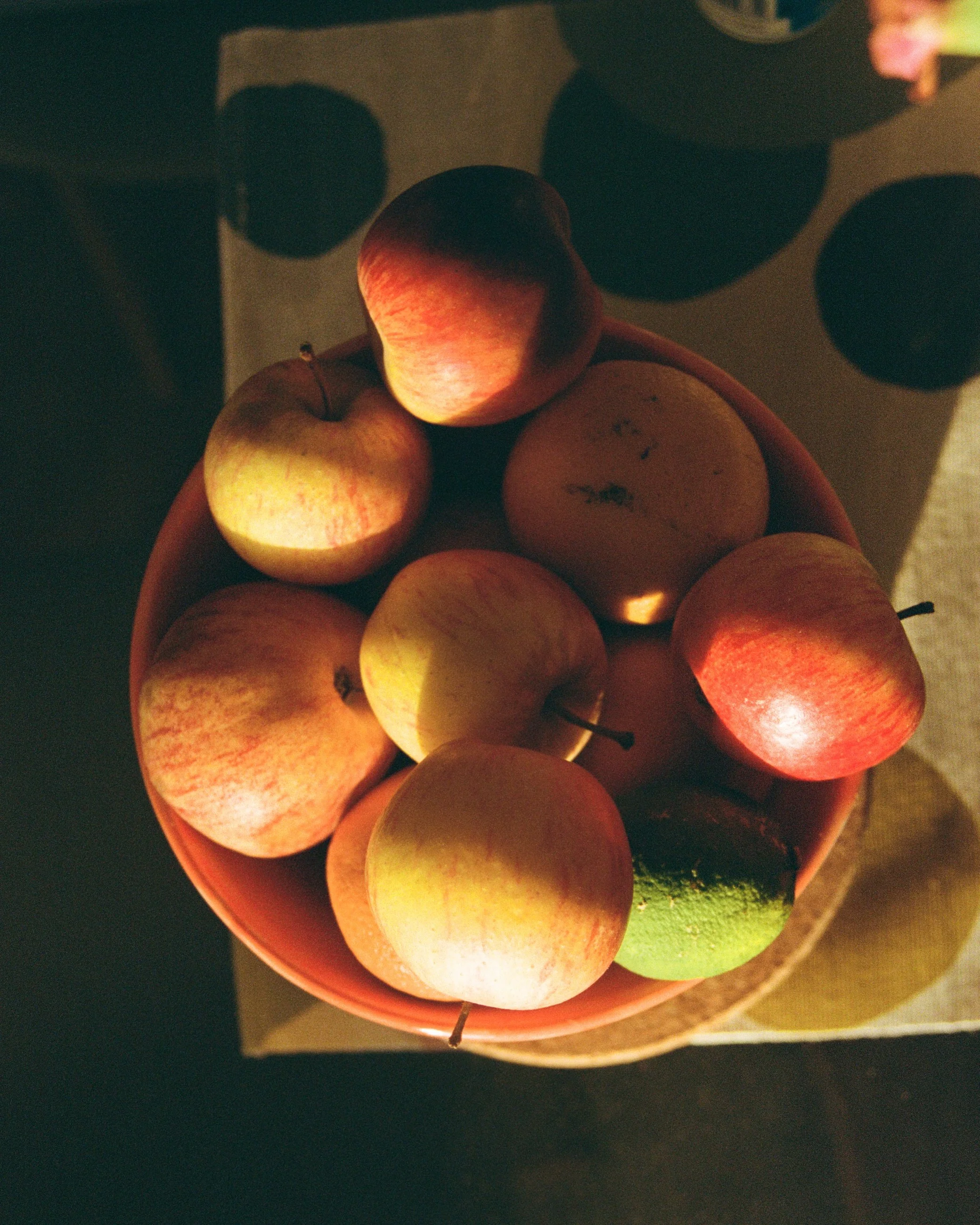 A bowl of mixed apples and at least one lime on a table, with shadows cast on the surface.