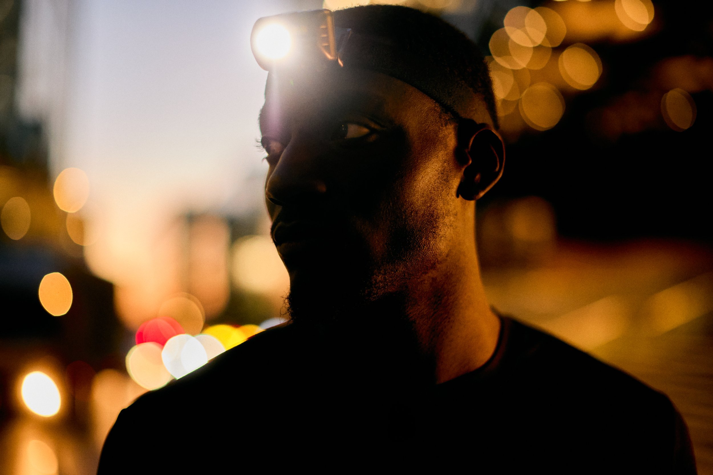 A silhouette of a man wearing a headlamp at dusk with city lights in the background.