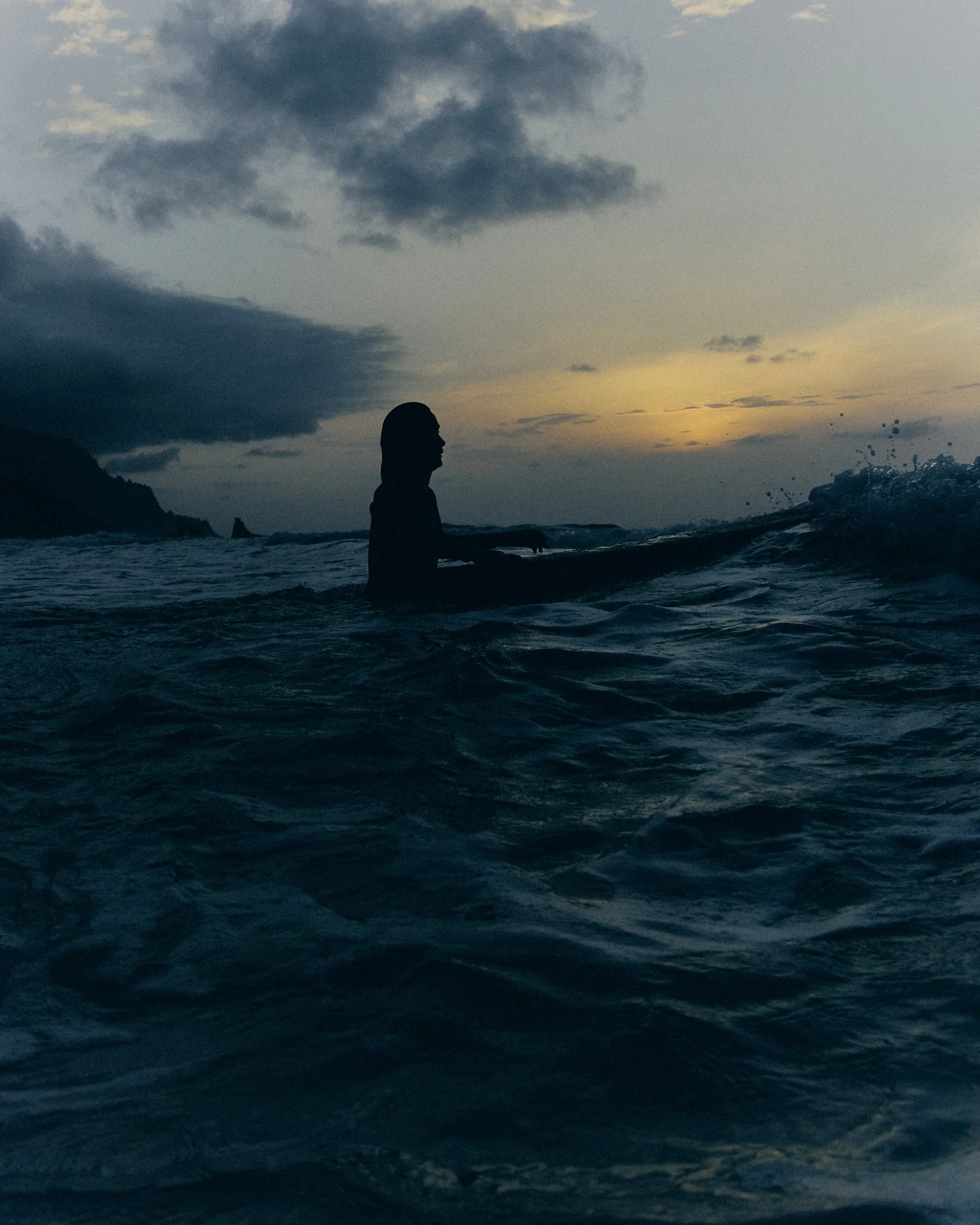 A person in the ocean during sunset or sunrise, with dark clouds in the sky and a distant landmass on the horizon.