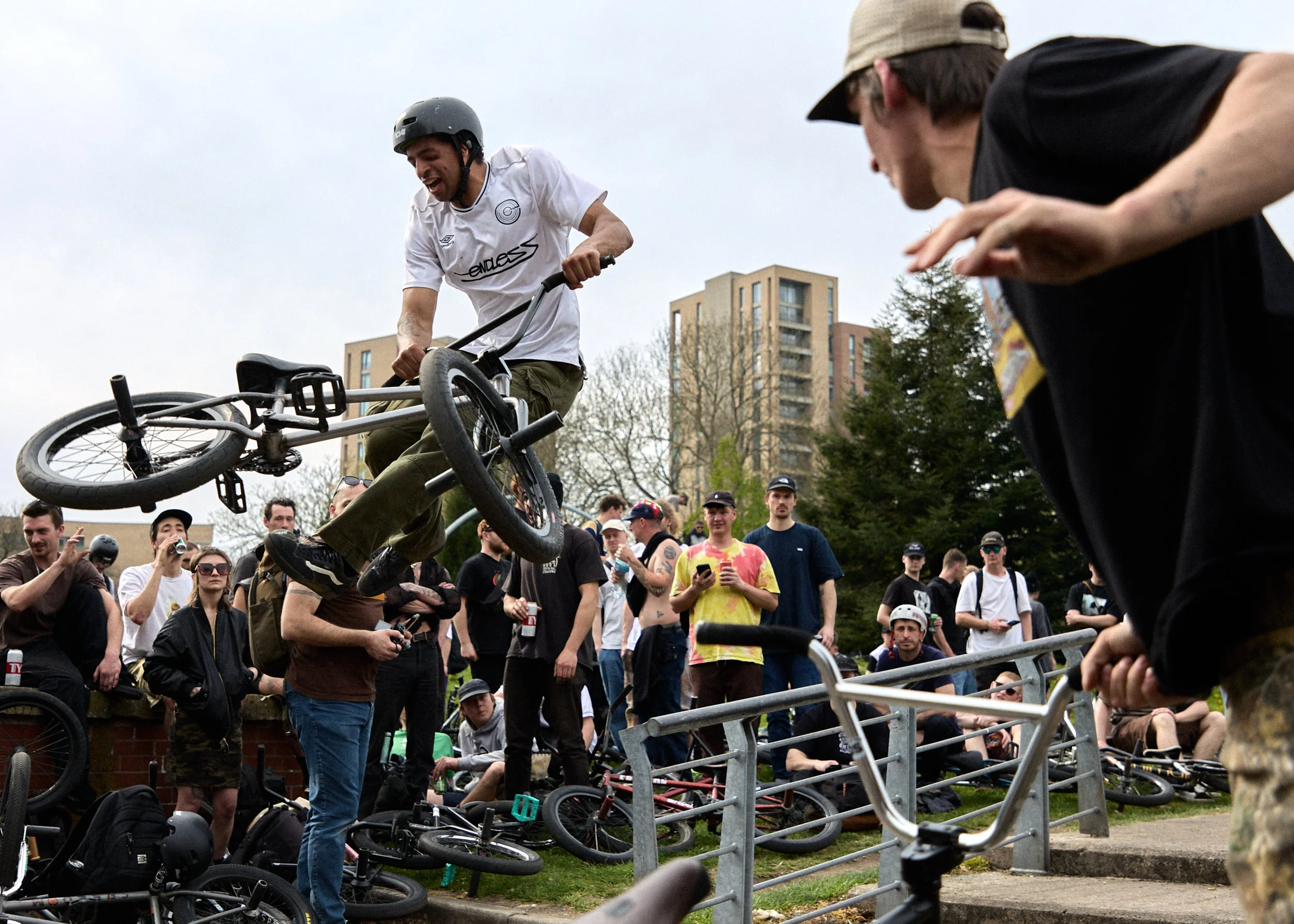 A young man wearing a helmet performs a BMX trick mid-air at a skatepark, with spectators watching and taking photos behind a metal railing.