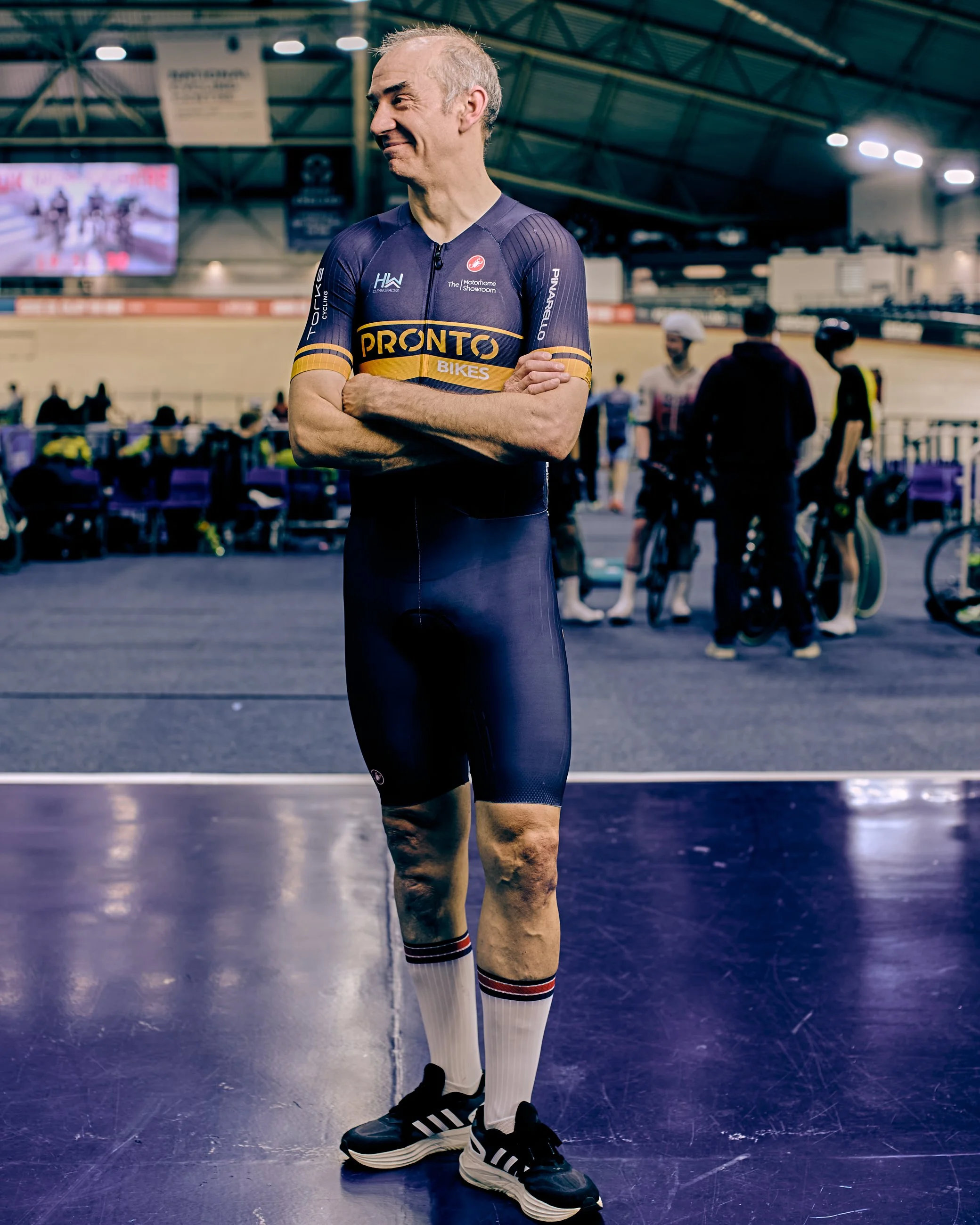 A man in a Pronto Bikes cycling uniform standing with his arms crossed inside a velodrome, with other cyclists and spectators in the background.