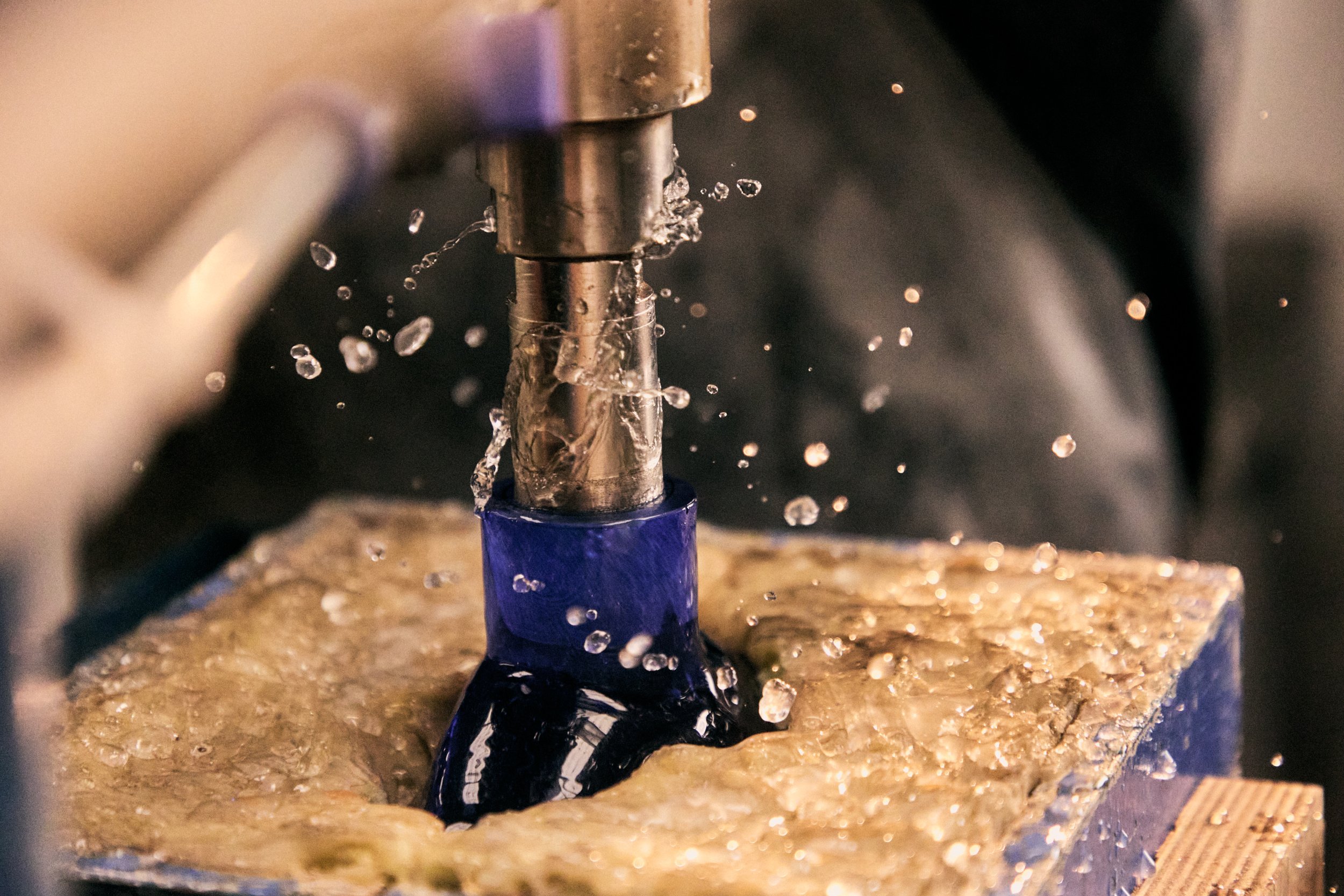 Close-up of a CNC machining process where a drill bit is cutting into a dark material, with water or coolant splashing around to reduce heat and remove debris.