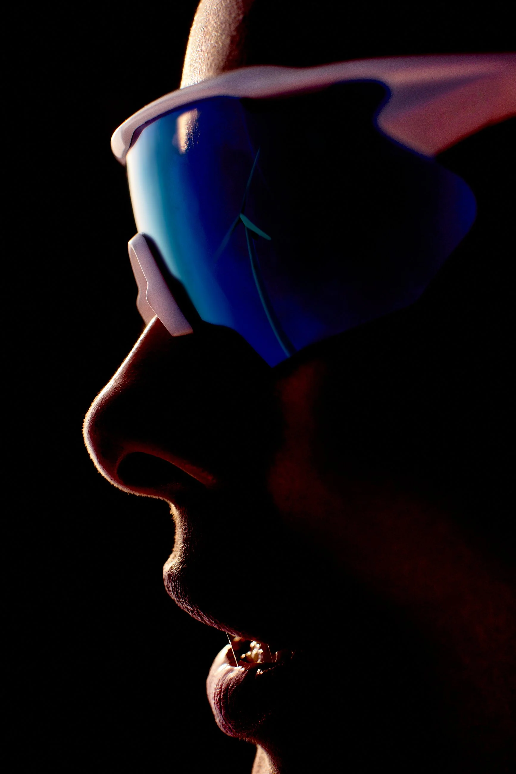 Close-up profile of a person wearing sunglasses with reflective blue lenses, illuminated from the side against a dark background.