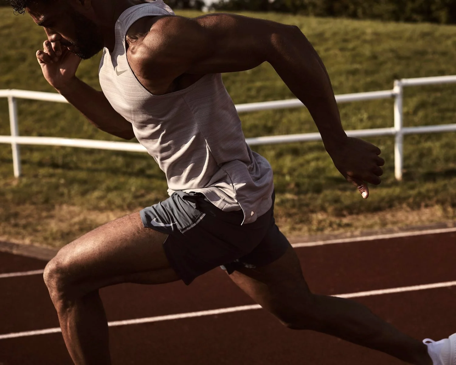 A male sprinter in a white tank top and black shorts running on a track outdoors.