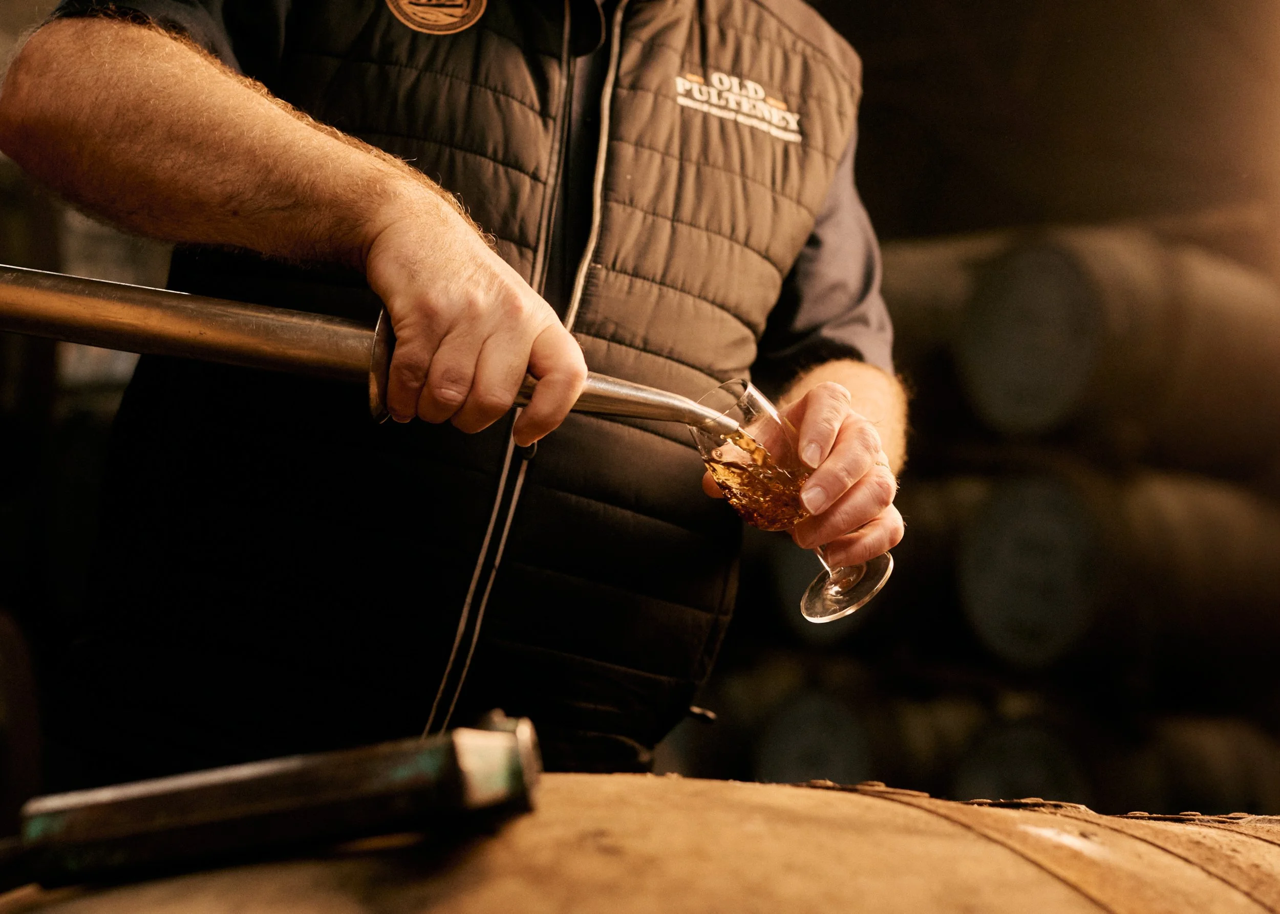 Person pouring whiskey into a glass from a metal funnel in a dimly lit setting with wooden barrels in the background.