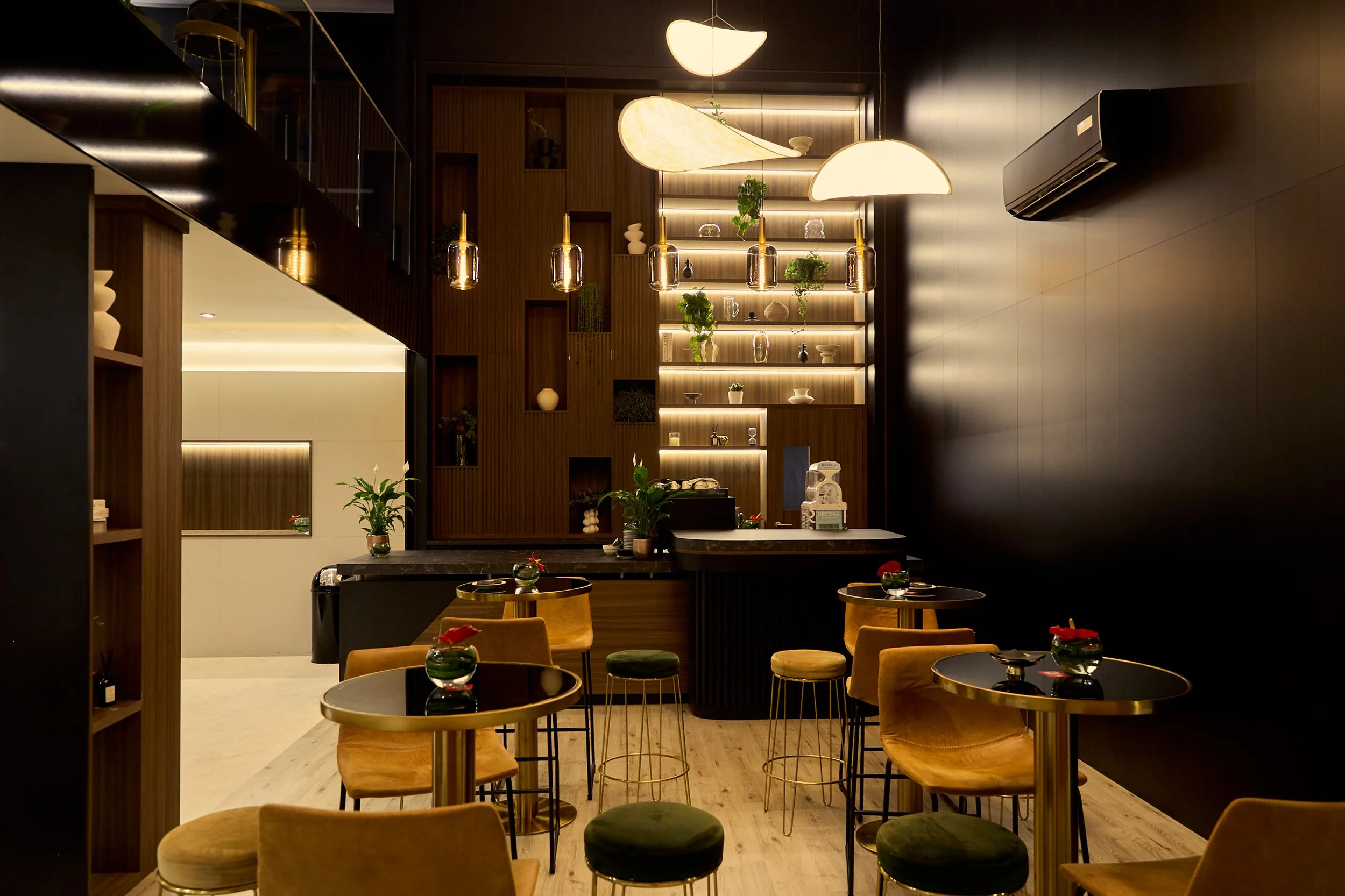 Interior of a modern, dimly-lit cafe with wooden and black walls, gold-accented furniture, and decorative lighting, featuring tables set with small green plants.