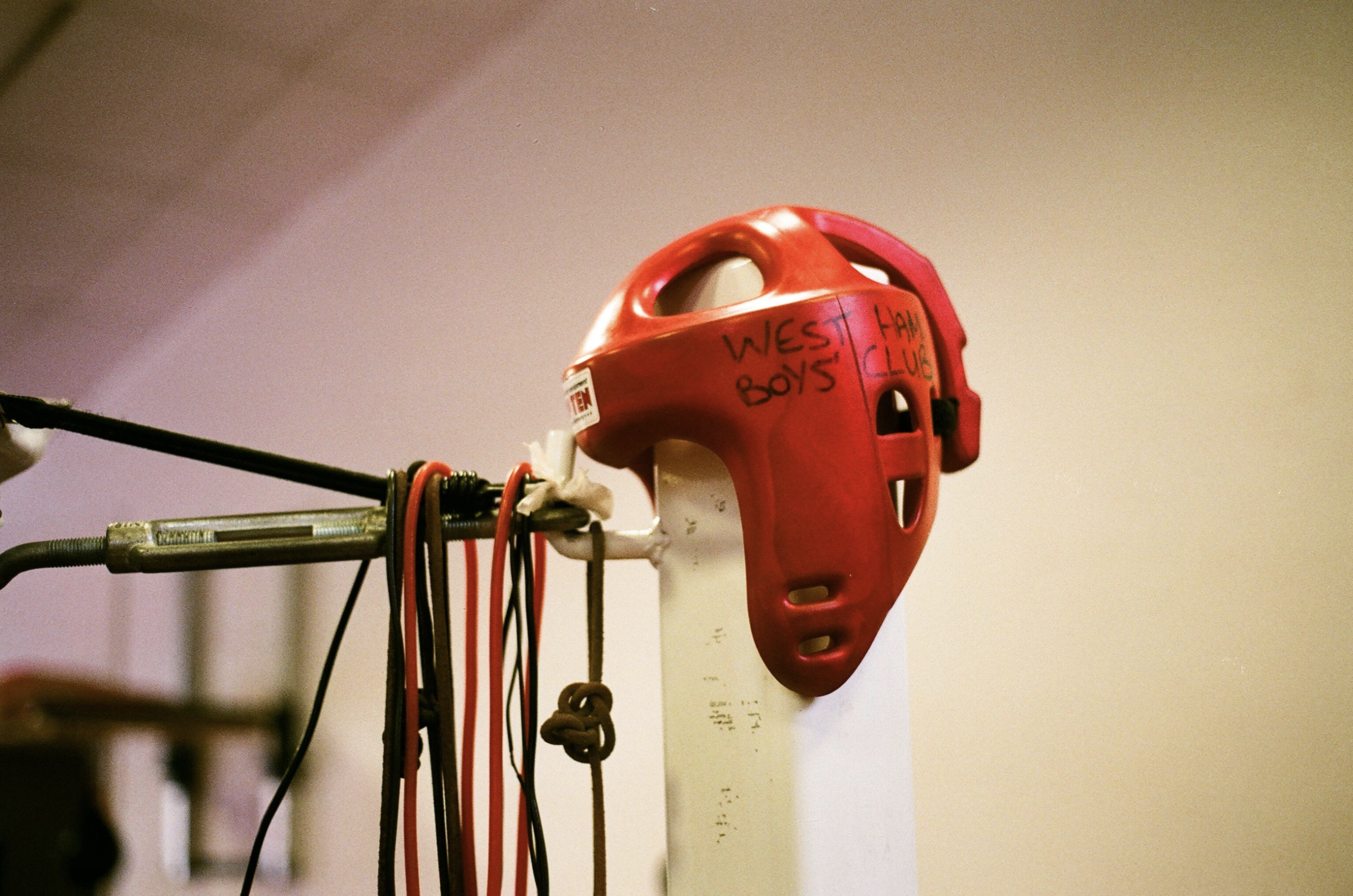 A red and white sports helmet with handwritten text reading 'West Ham Boys Club' hangs on a rack surrounded by various cords and straps.