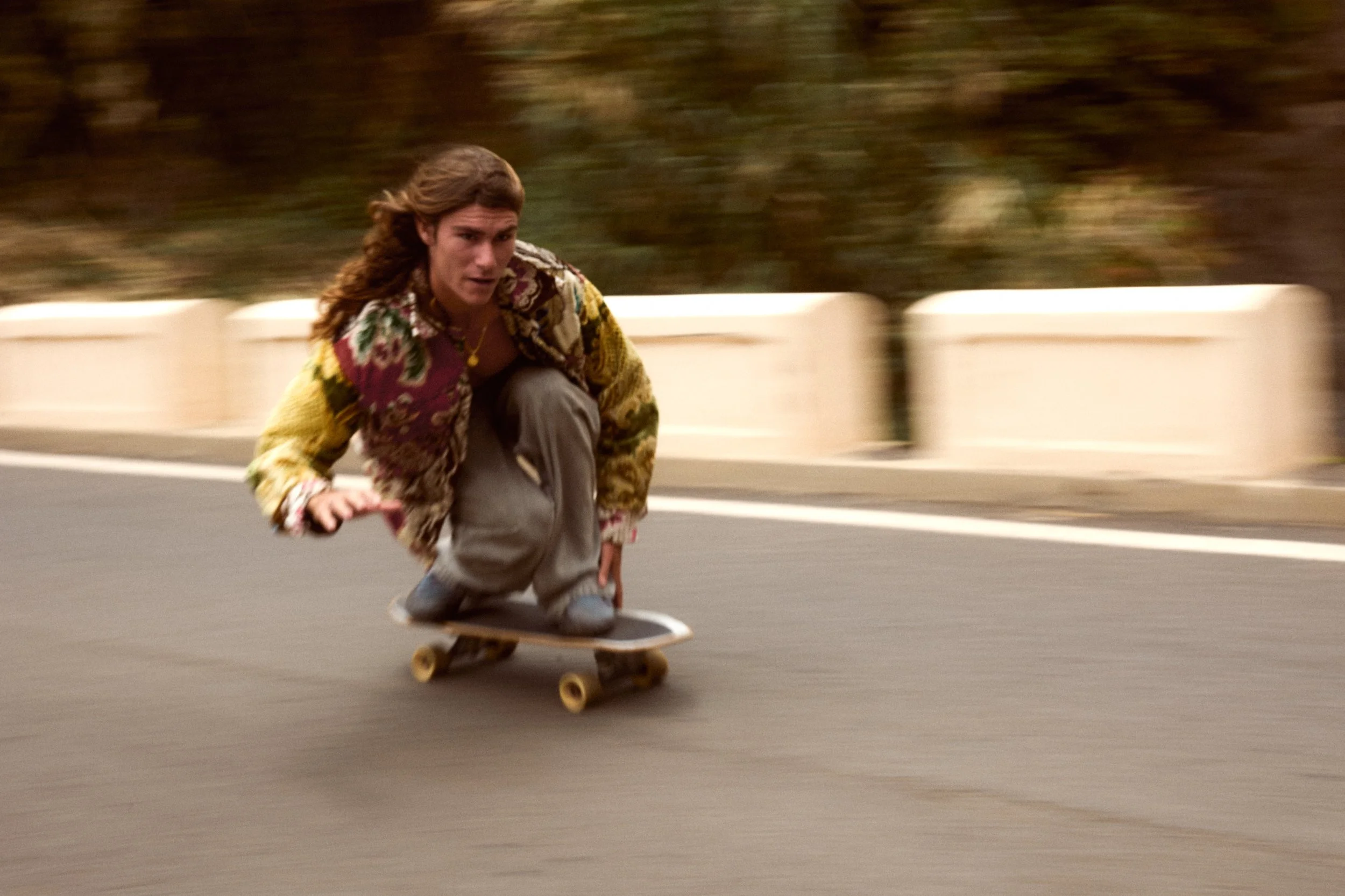 A person with long hair riding a skateboard on a road, wearing a colorful jacket and gray pants, with a background of trees and a barrier.