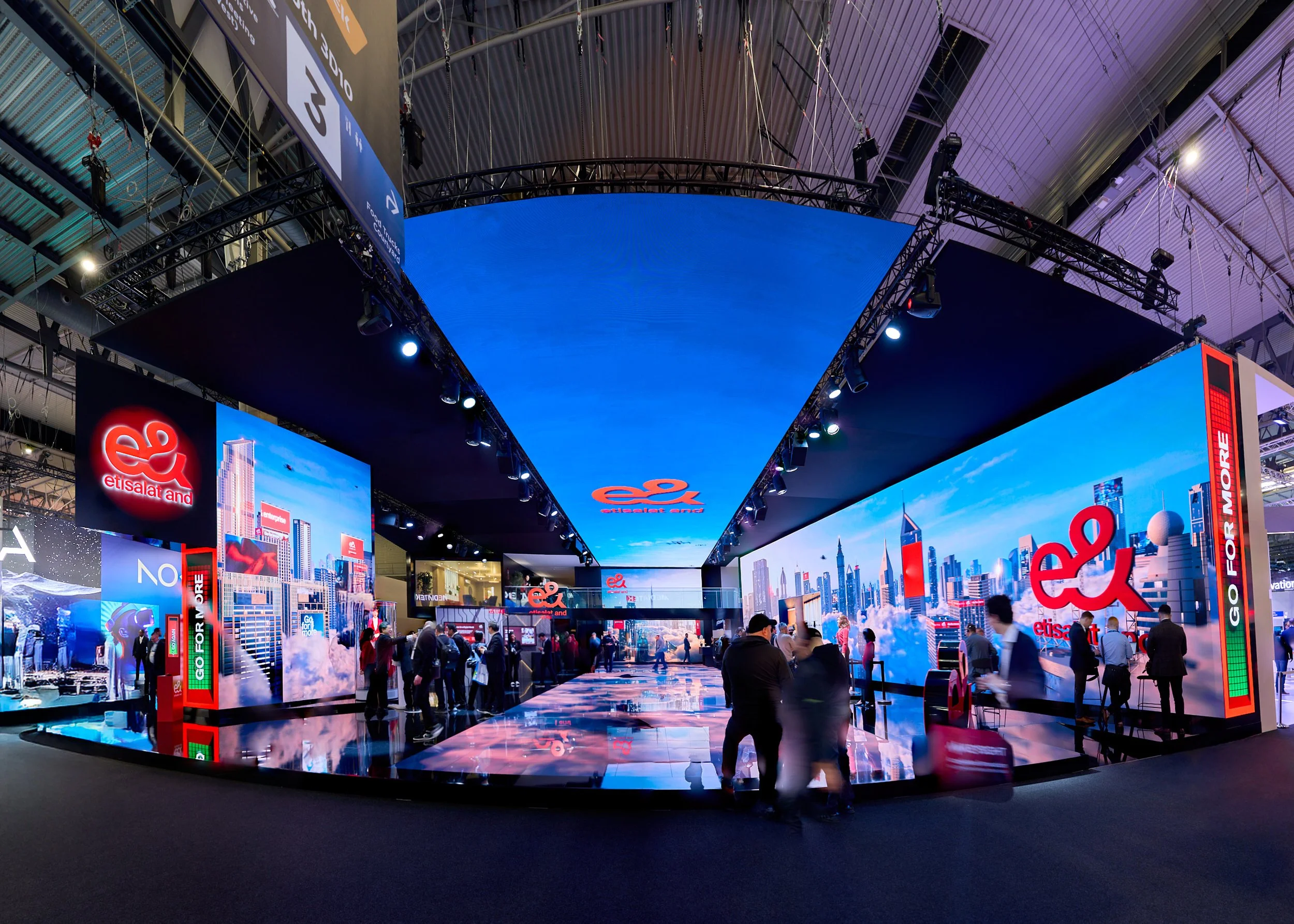 Indoor exhibition booth with digital displays and the logo of Etisalat and e&, showcasing a modern cityscape with skyscrapers, visitors walking around, and a blue ceiling overhead.