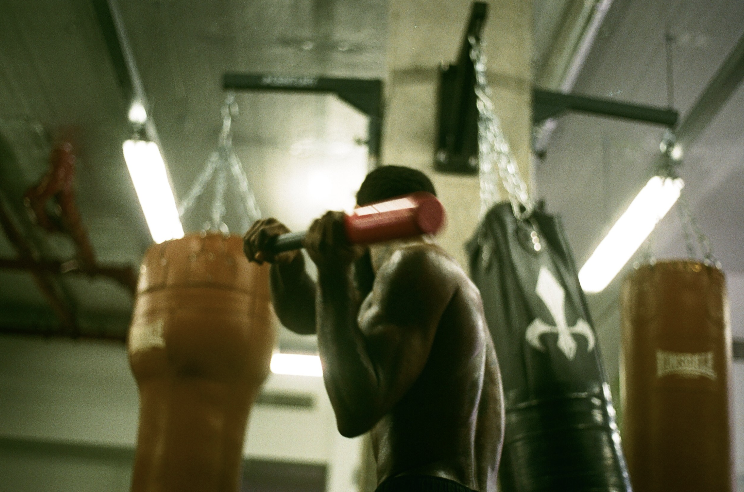 A muscular man lifts a barbell in a gym with punching bags hanging in the background.