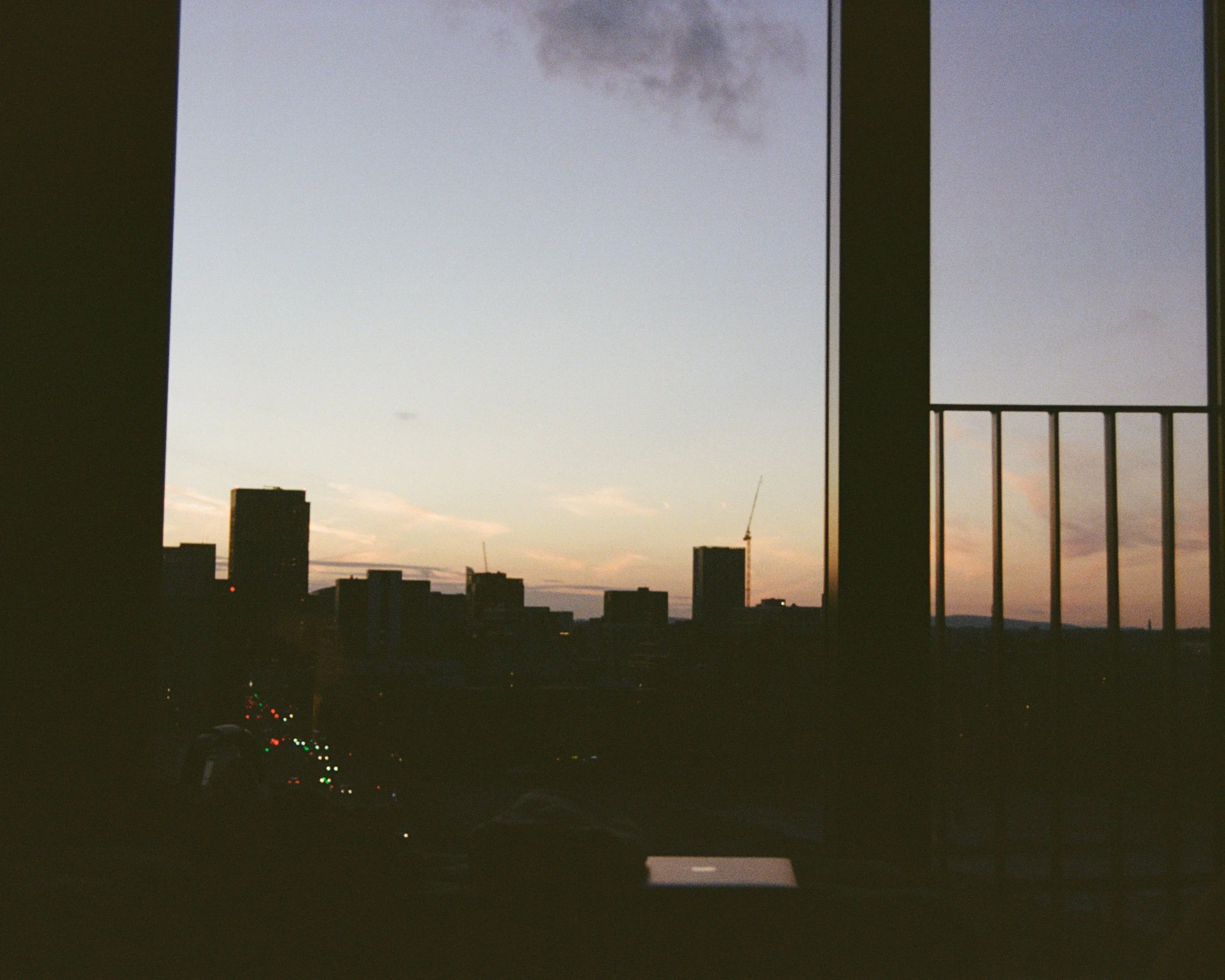 City skyline silhouette during sunset, viewed through a window with vertical bars on the right side, with some cloud cover in the sky.