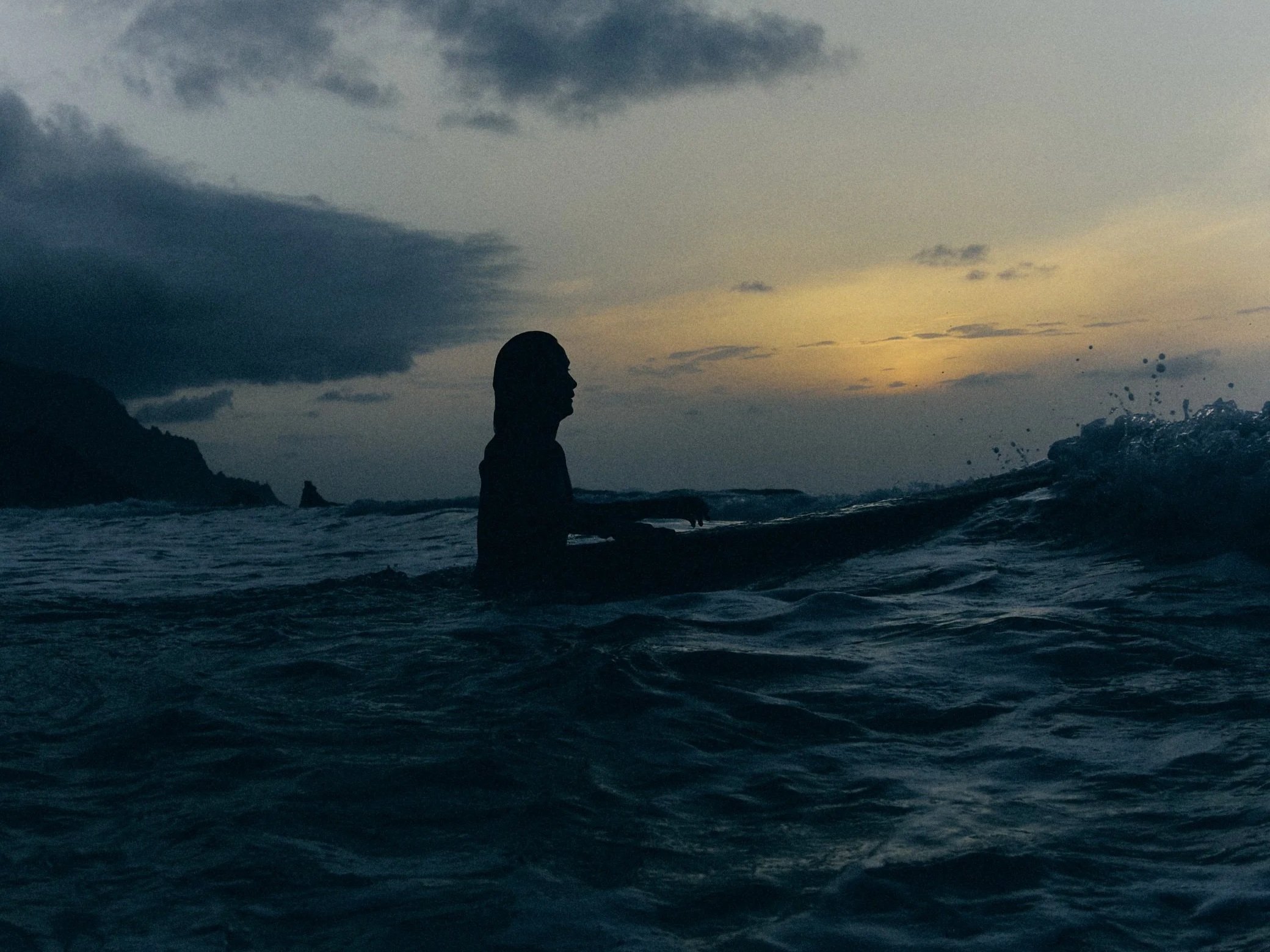 Silhouette of a person sitting in the water during sunset or dusk, with clouds in the sky and a distant rocky shoreline.