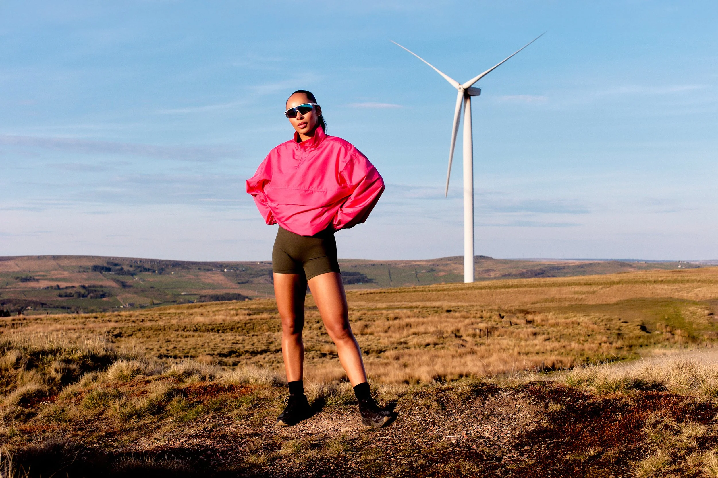 A woman wearing sunglasses, a pink jacket, black shorts, and boots standing on a grassy field with a wind turbine in the background.