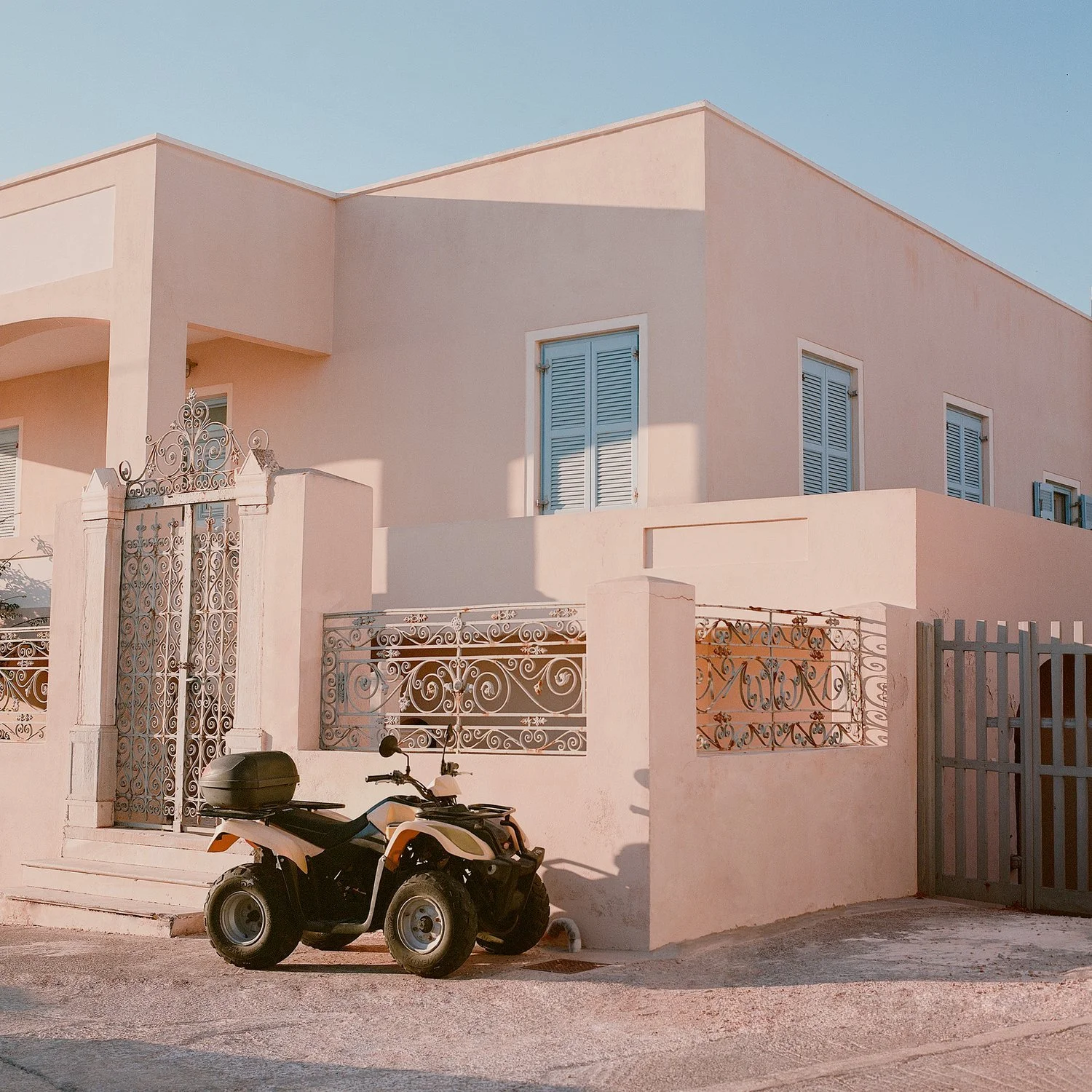 A pink multi-story house with white shutters on its windows and decorative iron gates at the entrance, with a black quad bike parked outside.