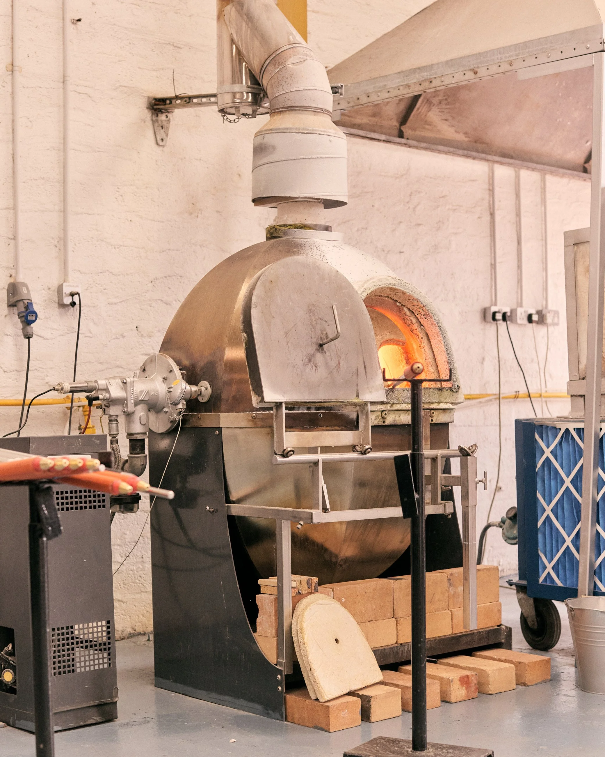 A commercial pizza oven with a rounded stainless steel exterior, open door revealing glowing interior fire, and bricks around it. There are various pipes and equipment attached, with a small stack of bricks and a circular stone piece in front.