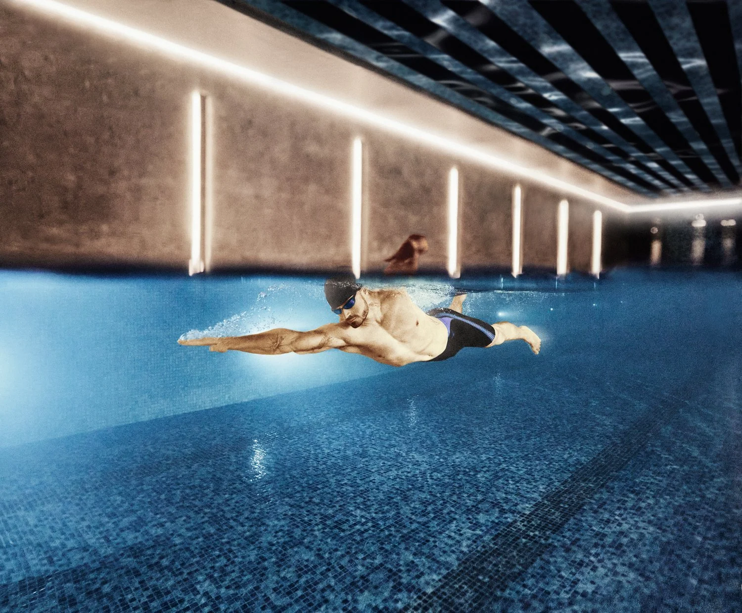 A male swimmer in black shorts with blue accents, wearing goggles, doing a front crawl stroke in an indoor swimming pool.