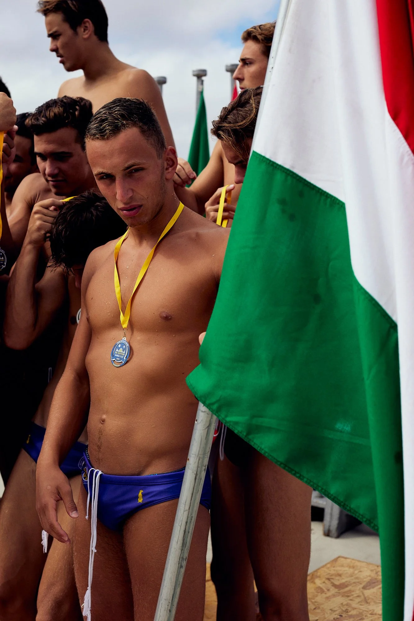 Young male swimmer with a medal around his neck, wearing blue swim trunks, among other swimmers, standing next to a large flag, outdoors during a sports event.