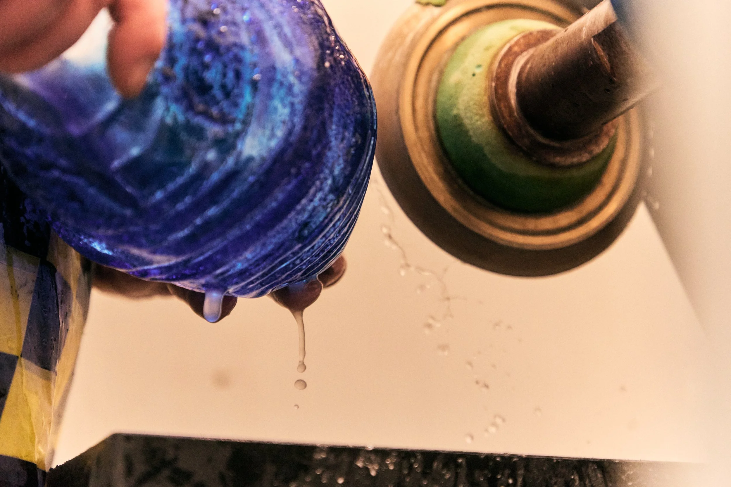 A person pouring water from a blue glass pitcher into a wooden container, with water spilling out onto a white surface.