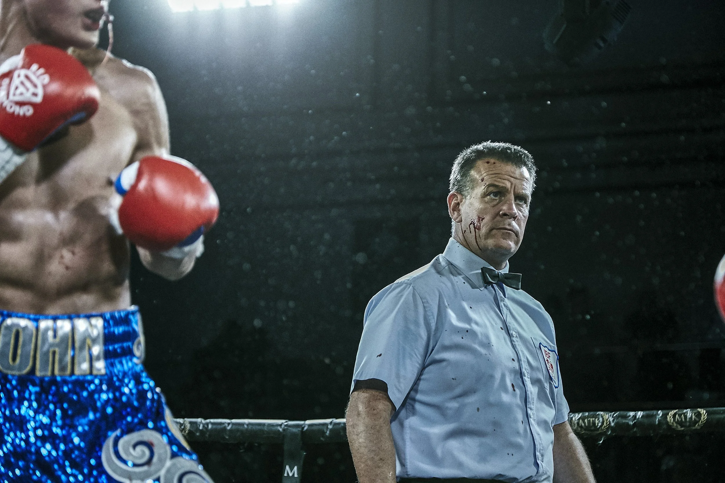 Boxer wearing red gloves and blue shorts with gold lettering, standing in a boxing ring, with a referee standing nearby with blood on his face and a serious expression.