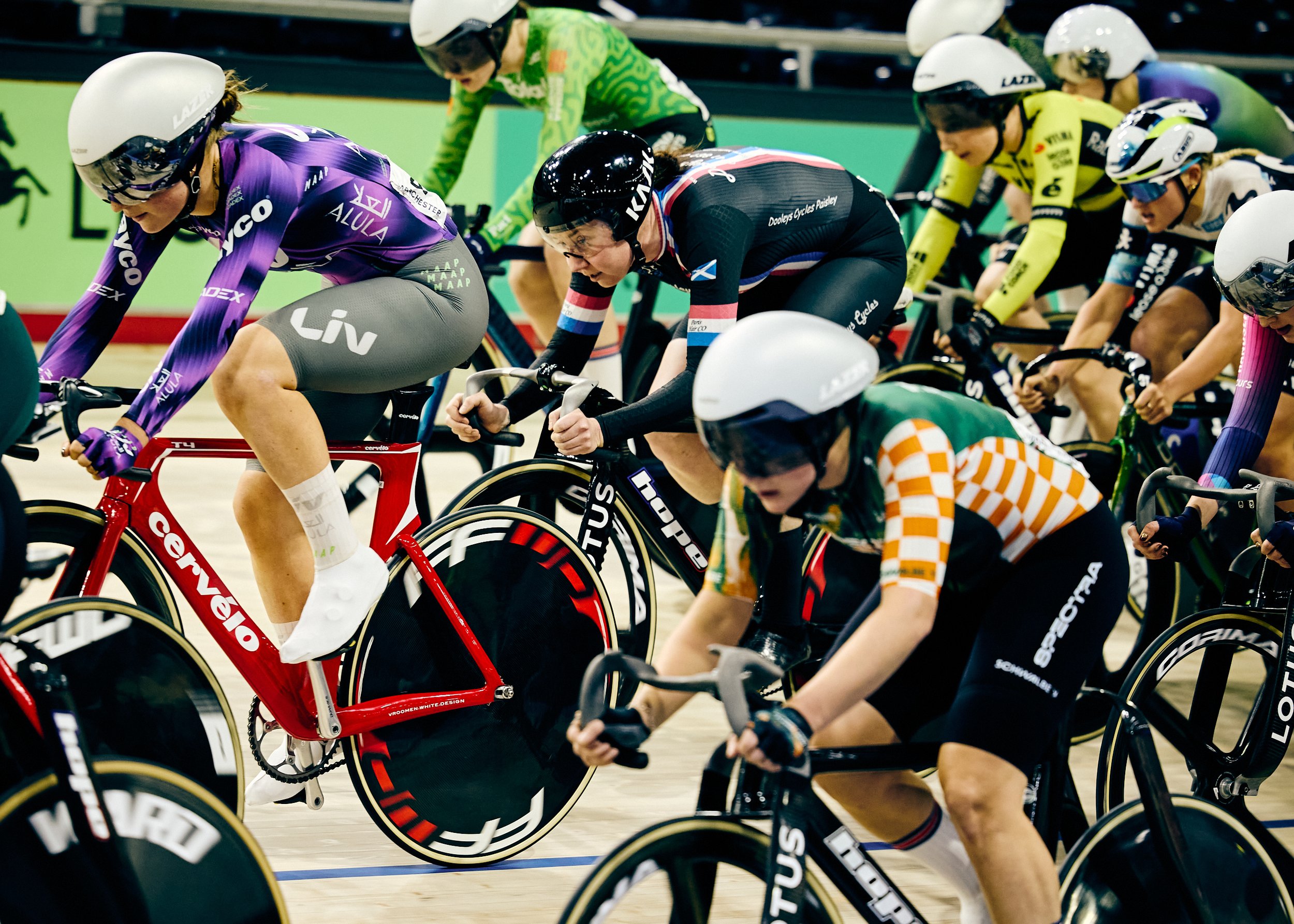 Multiple cyclists racing on an indoor velodrome, wearing helmets and colorful racing outfits, with streamlined bikes, in a competitive track cycling event.