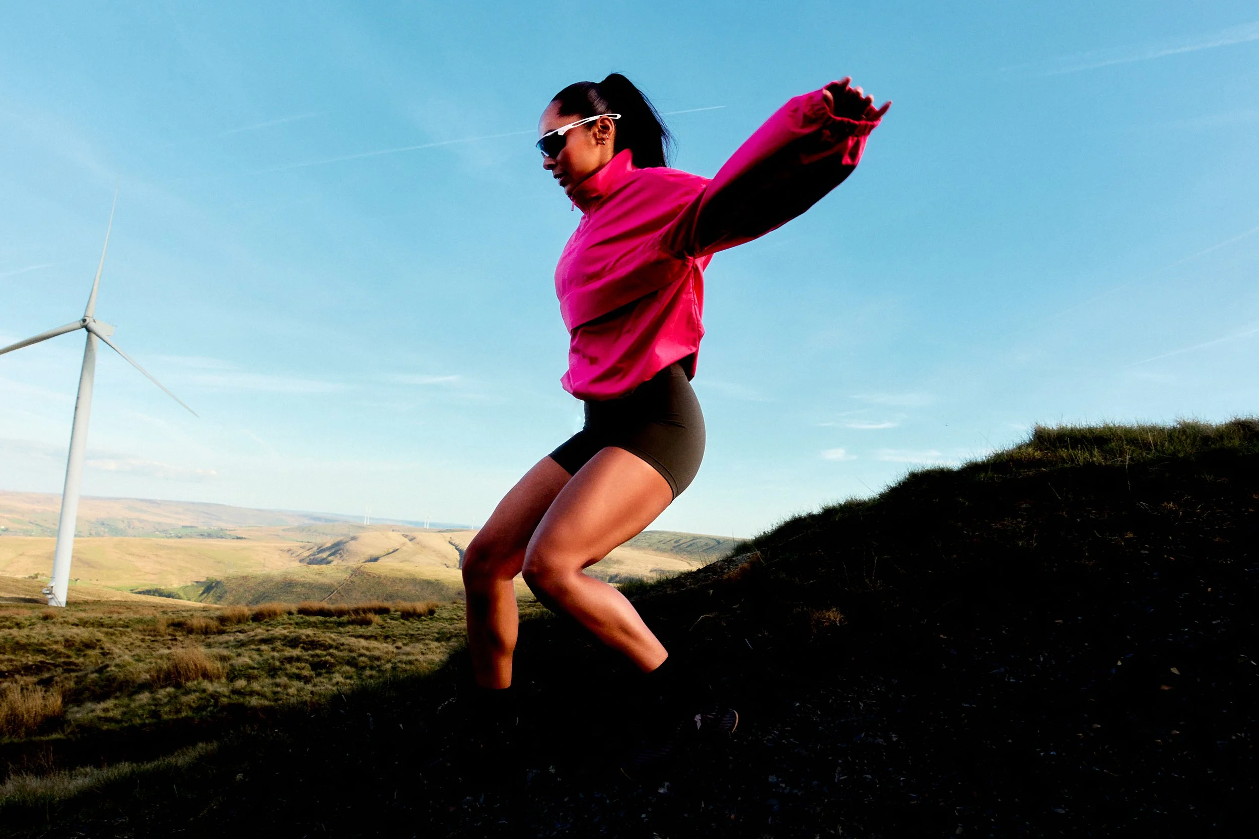 A woman wearing a pink jacket, black shorts, and sunglasses is jumping in a hilly outdoor landscape with wind turbines in the background under a clear blue sky.