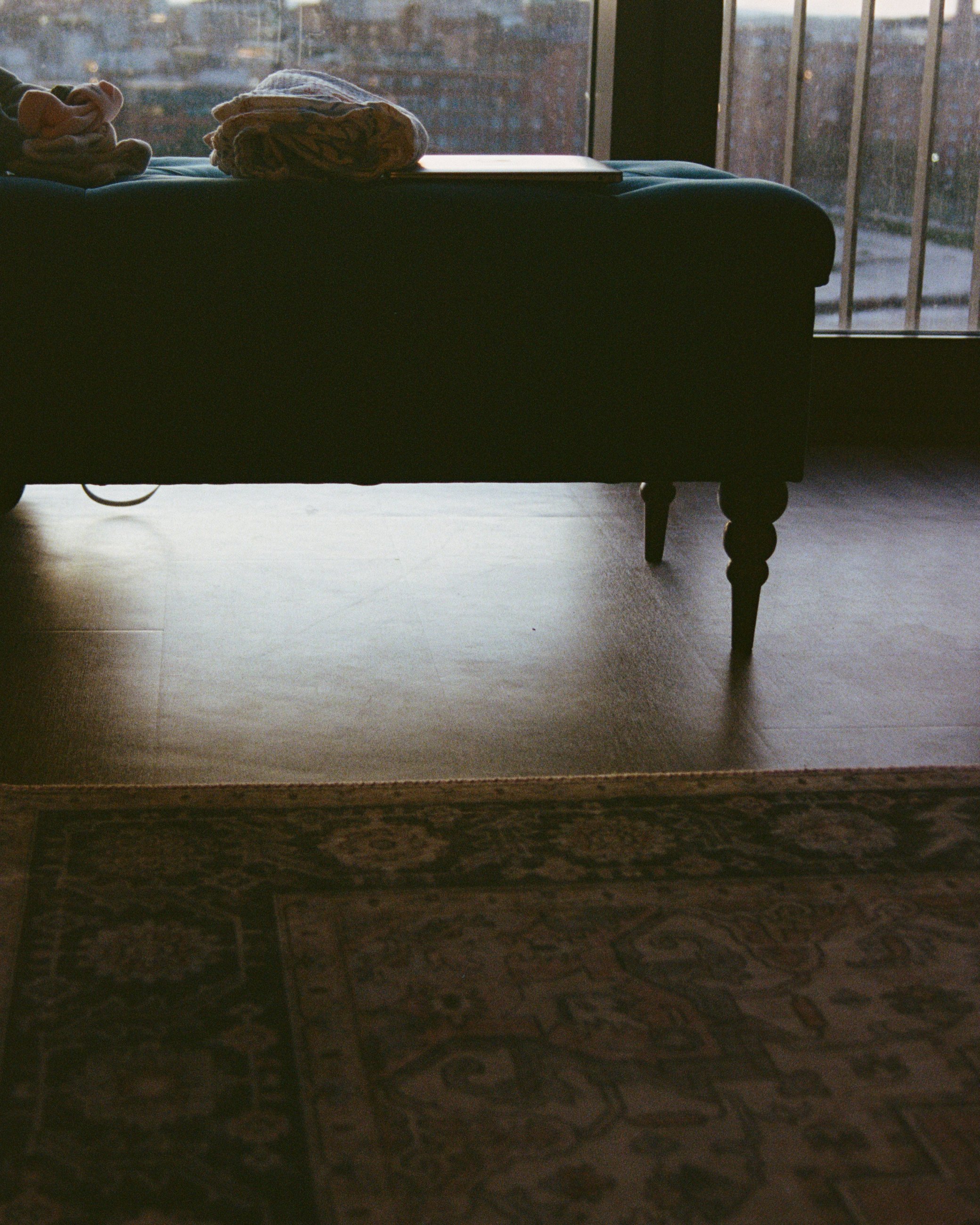 A silhouette of a dark-colored sofa with wooden legs, placed on a wooden floor near large windows, with a patterned rug partially visible at the bottom of the image.