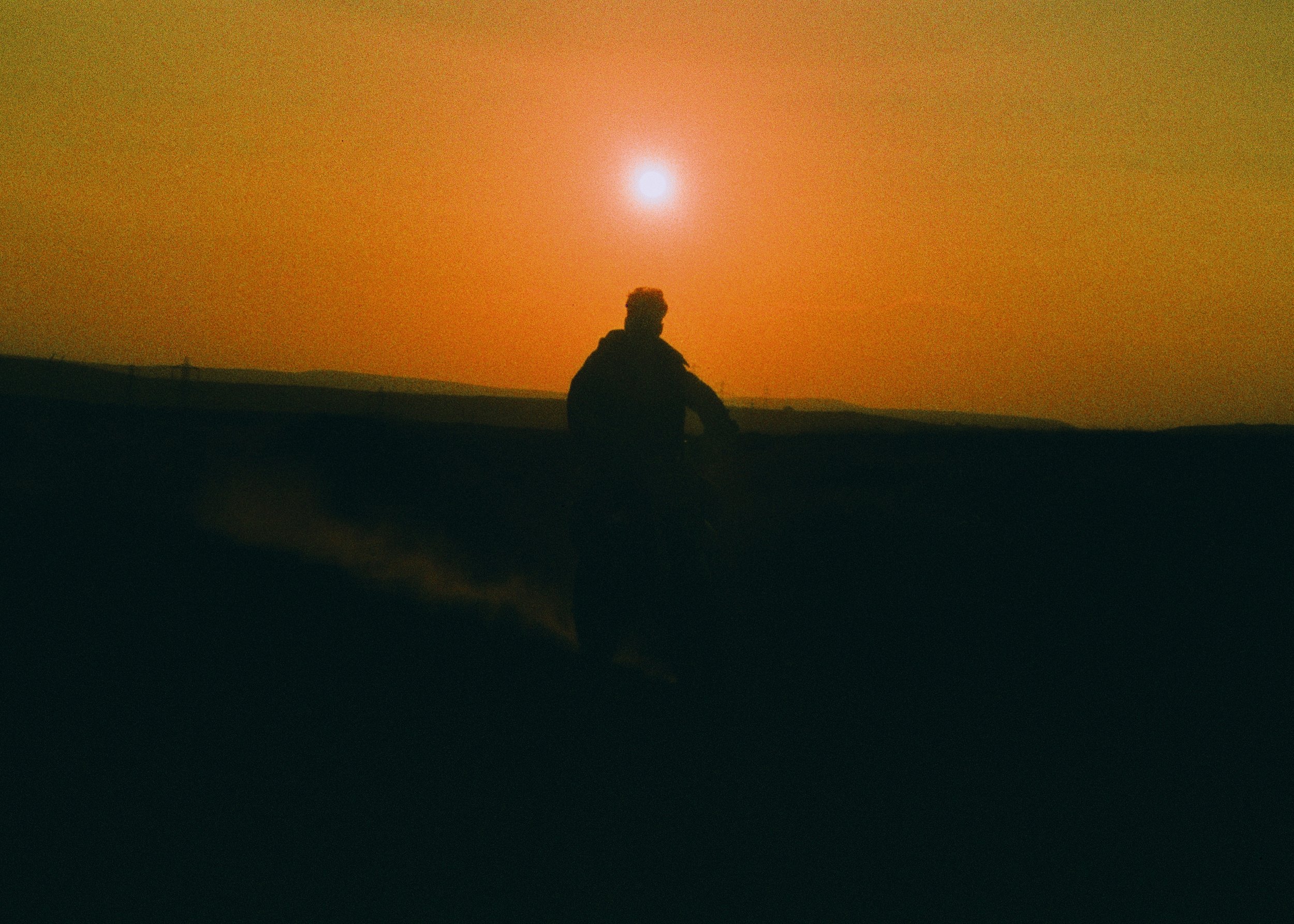 Silhouette of a person sitting on a hill at sunset or sunrise with a bright sun in the sky and an orange hue over the landscape.