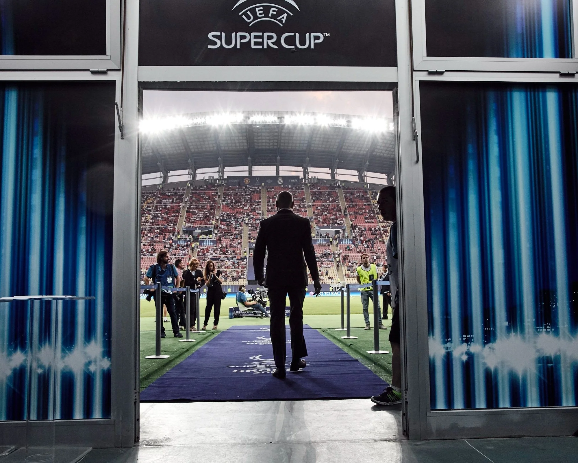 People walking into a stadium for the UEFA Super Cup match, with a view of the field and audience in the background.