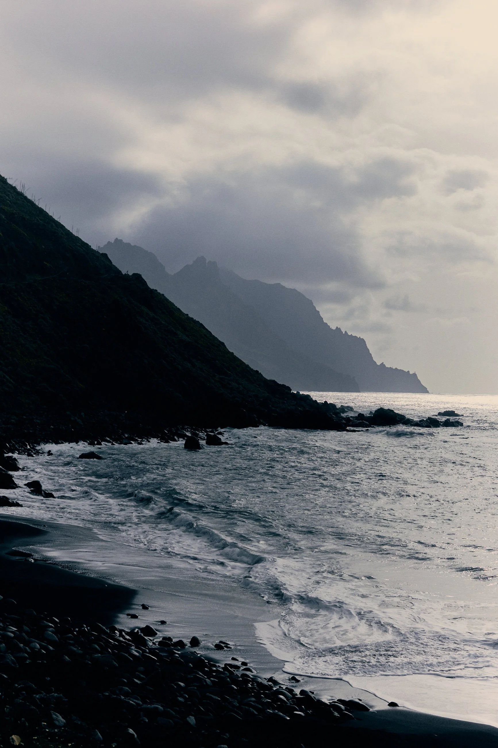 A rocky shoreline with waves crashing against the coast, steep green mountains in the background, and a cloudy sky overhead.