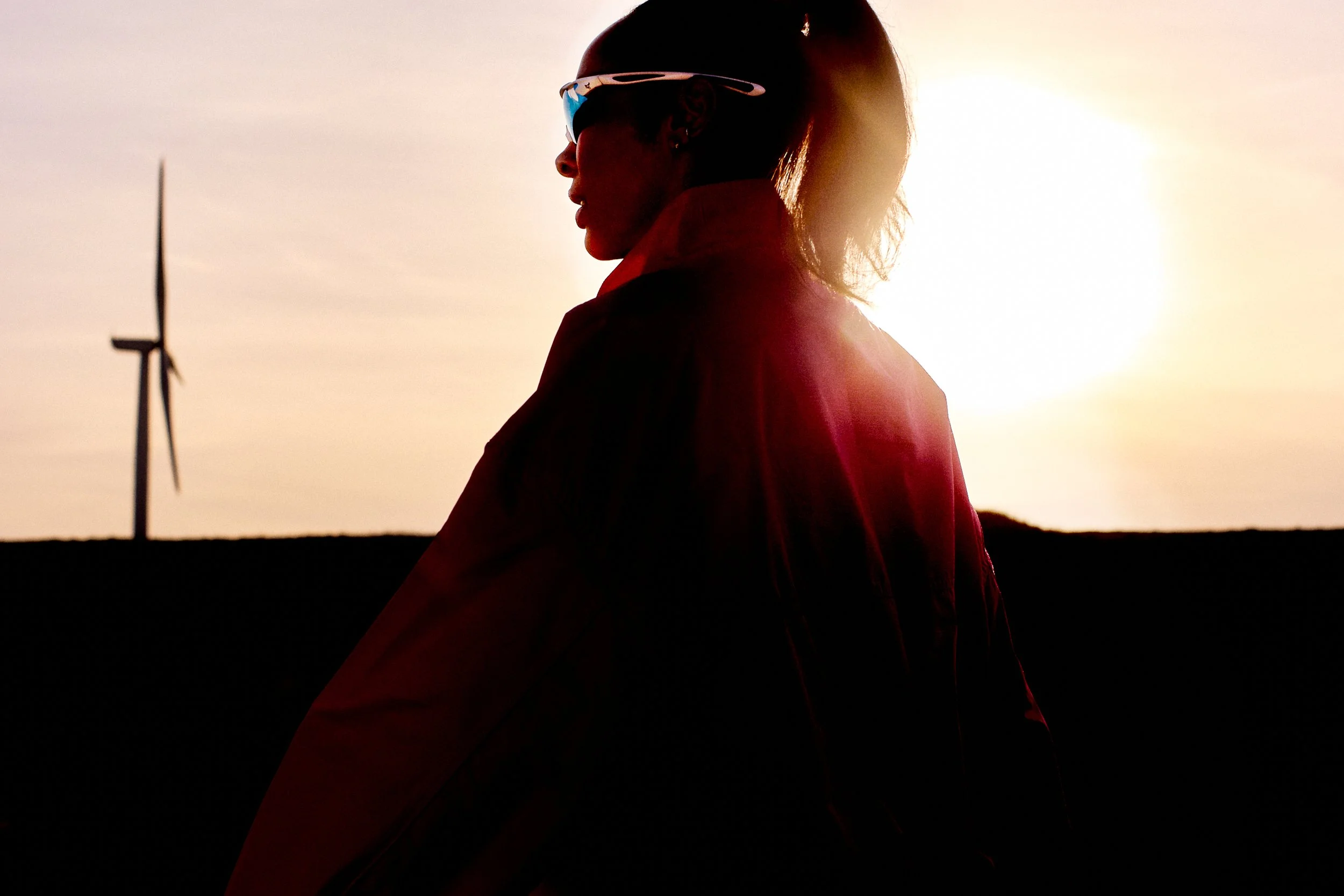 Silhouette of a woman wearing sunglasses, standing outdoors at sunset with a wind turbine in the background.