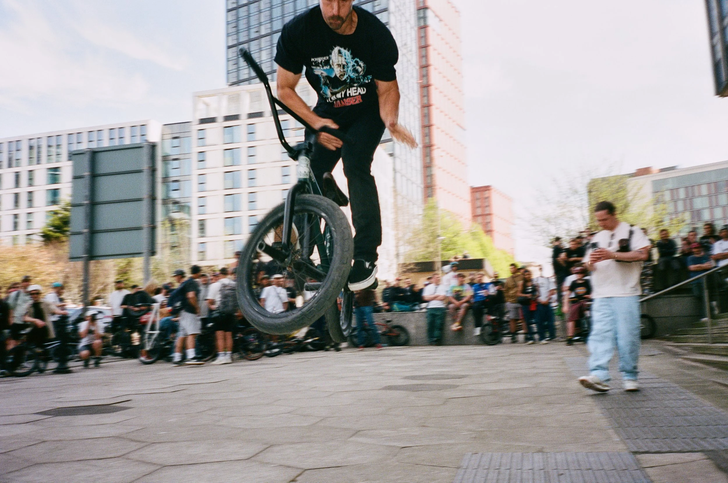 A man performing a BMX trick in an urban plaza with a crowd watching.