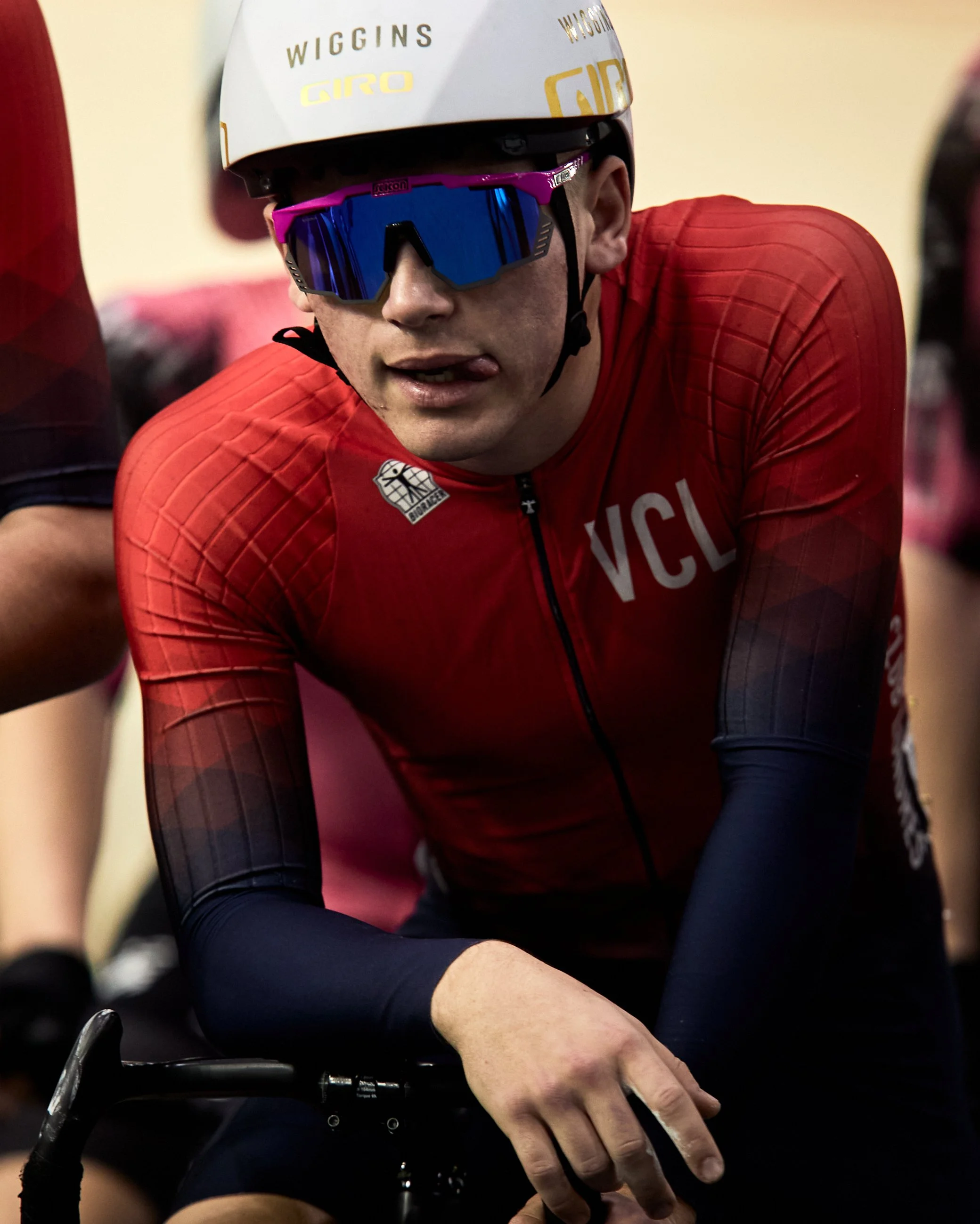 Cyclist wearing a white helmet with gold lettering, blue sunglasses with pink frame, and a red jersey with black sleeves, leaning forward on his bike.