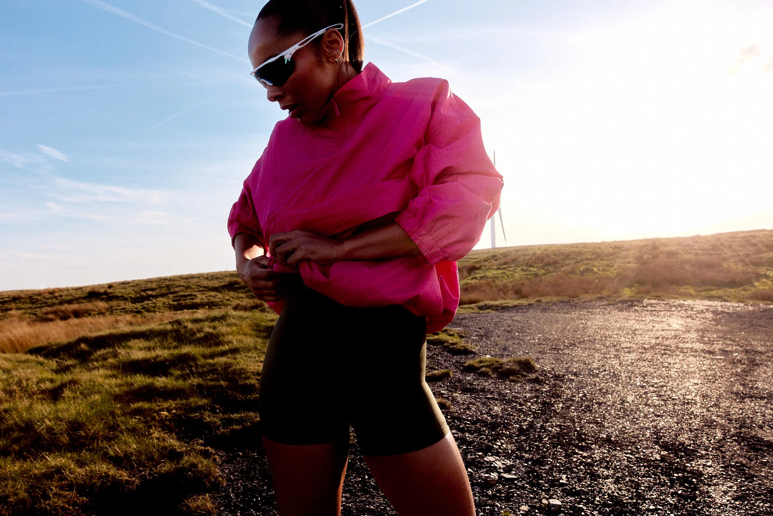 A woman wearing athletic clothing and sunglasses adjusting her pink jacket on a trail in a grassy, hilly landscape during sunset or sunrise.