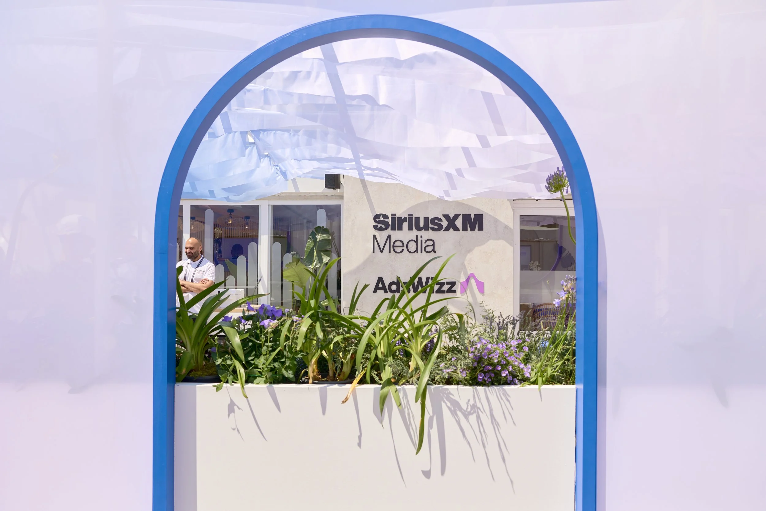 View through a white and blue arched opening to a garden with various green plants and purple flowers, with the SiriusXM Media and AdWizz logos on a building in the background.