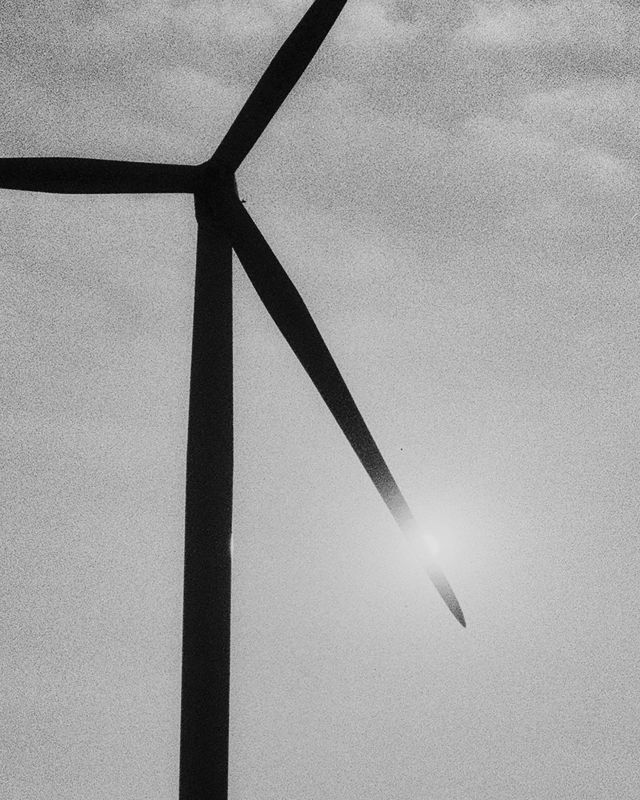 Black and white photograph of a wind turbine against the sky with the sun shining near the blades.