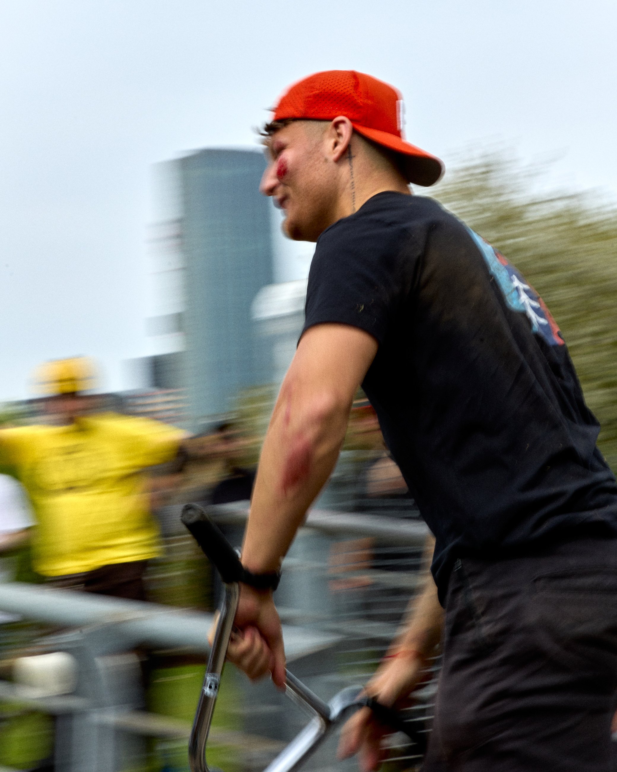 Young man with a red backwards cap and black t-shirt riding a BMX bike during a race or event, with the background showing blurred people and city buildings.
