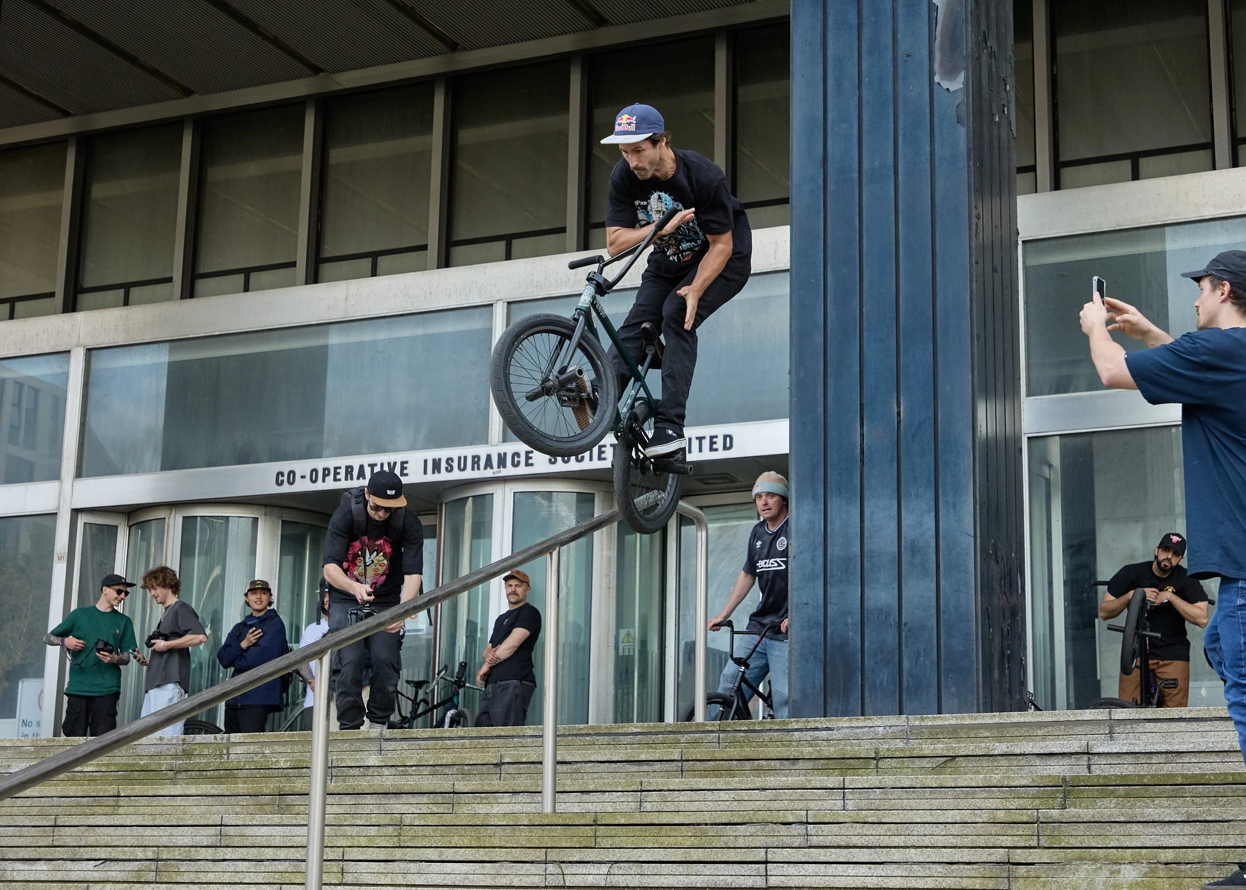 A BMX rider performs a trick on a rail outside a building while others watch and record. The building has a sign for the Co-Operative Insurance Society Limited.