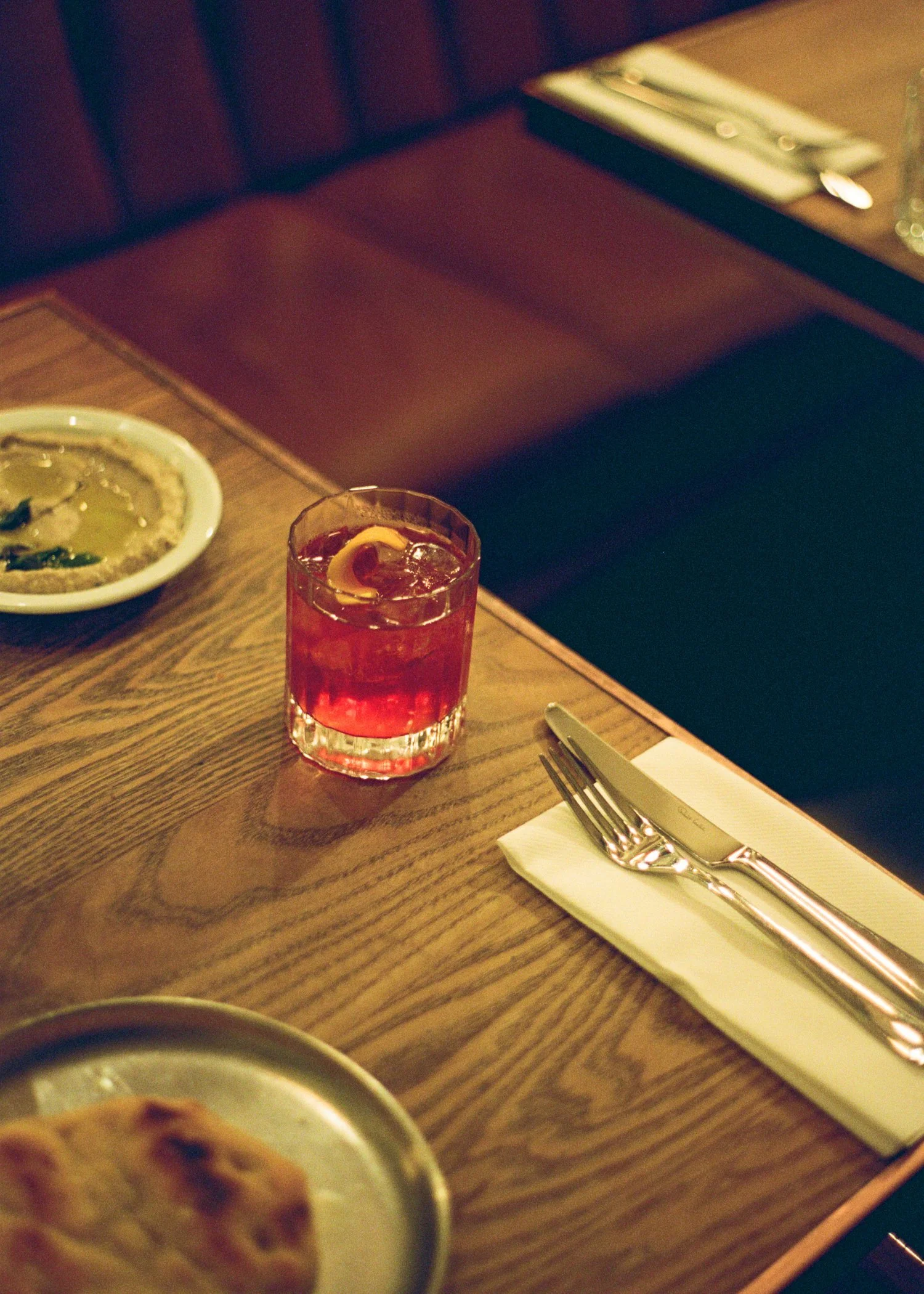 A wooden table with a red cocktail glass, a plate of food, a fork, and a knife on a napkin in a dimly lit restaurant.