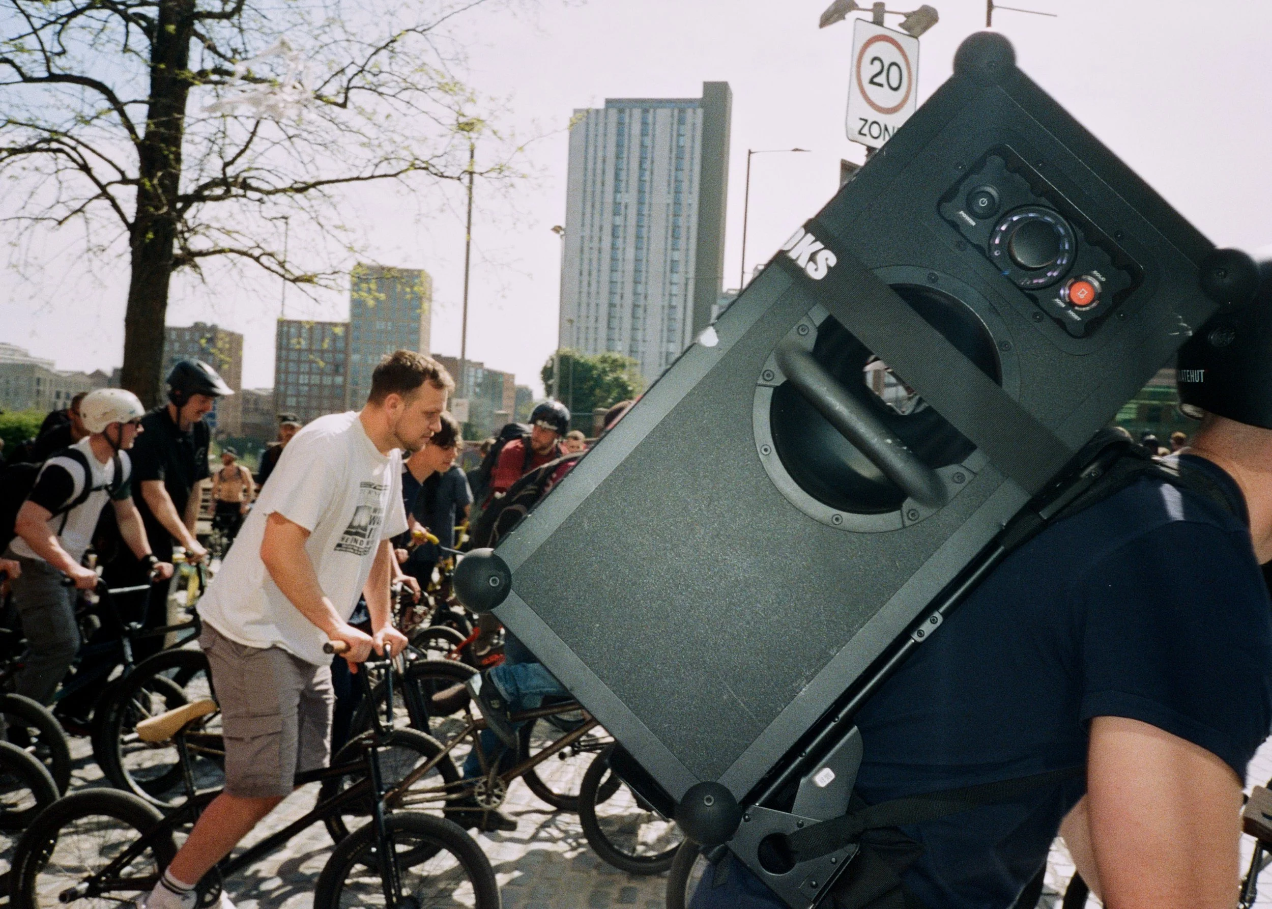 A man is riding a bicycle during a group bike ride event in an urban park, with a large speaker backpack on his back. The background features high-rise buildings and clear weather.